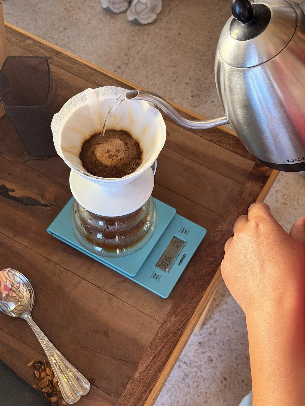Pouring hot water into a coffee filter with coffee grounds on a kitchen scale, preparing pour-over coffee on a wooden table.