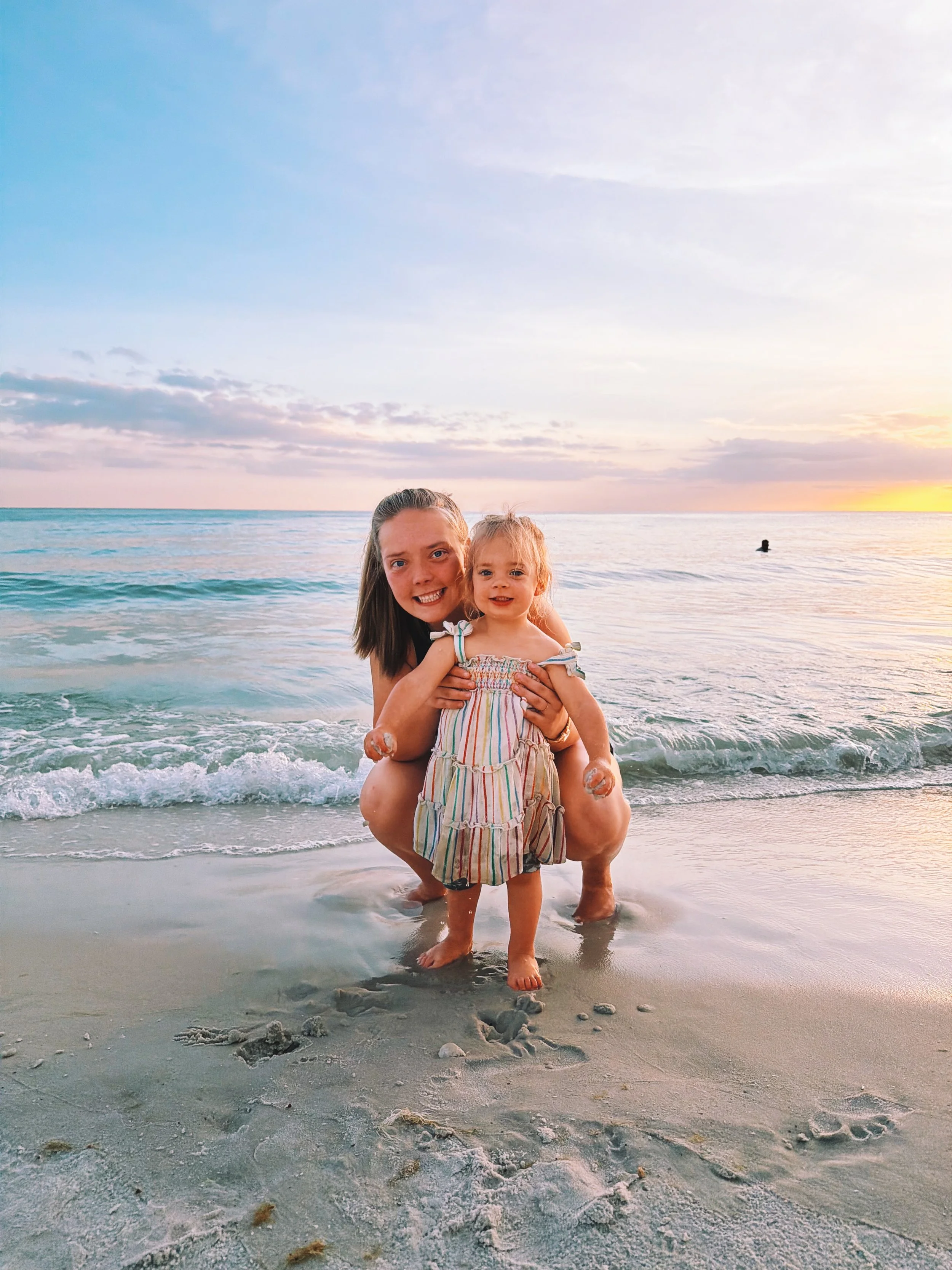 A woman holding a young girl standing in the shallow water at the beach during sunset with a person swimming in the background.