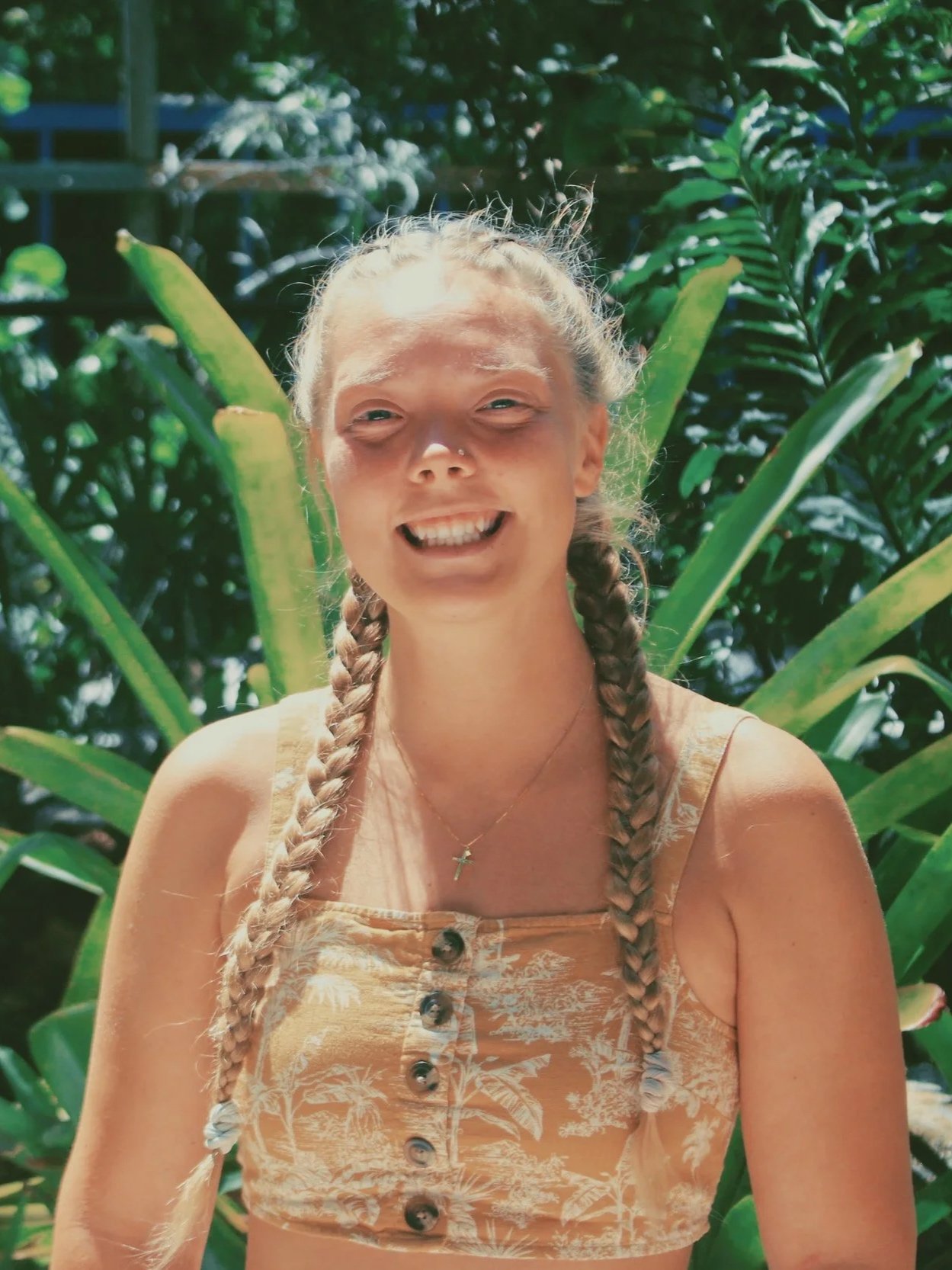 A young woman with braided hair smiling outdoors, surrounded by green tropical plants on a sunny day.