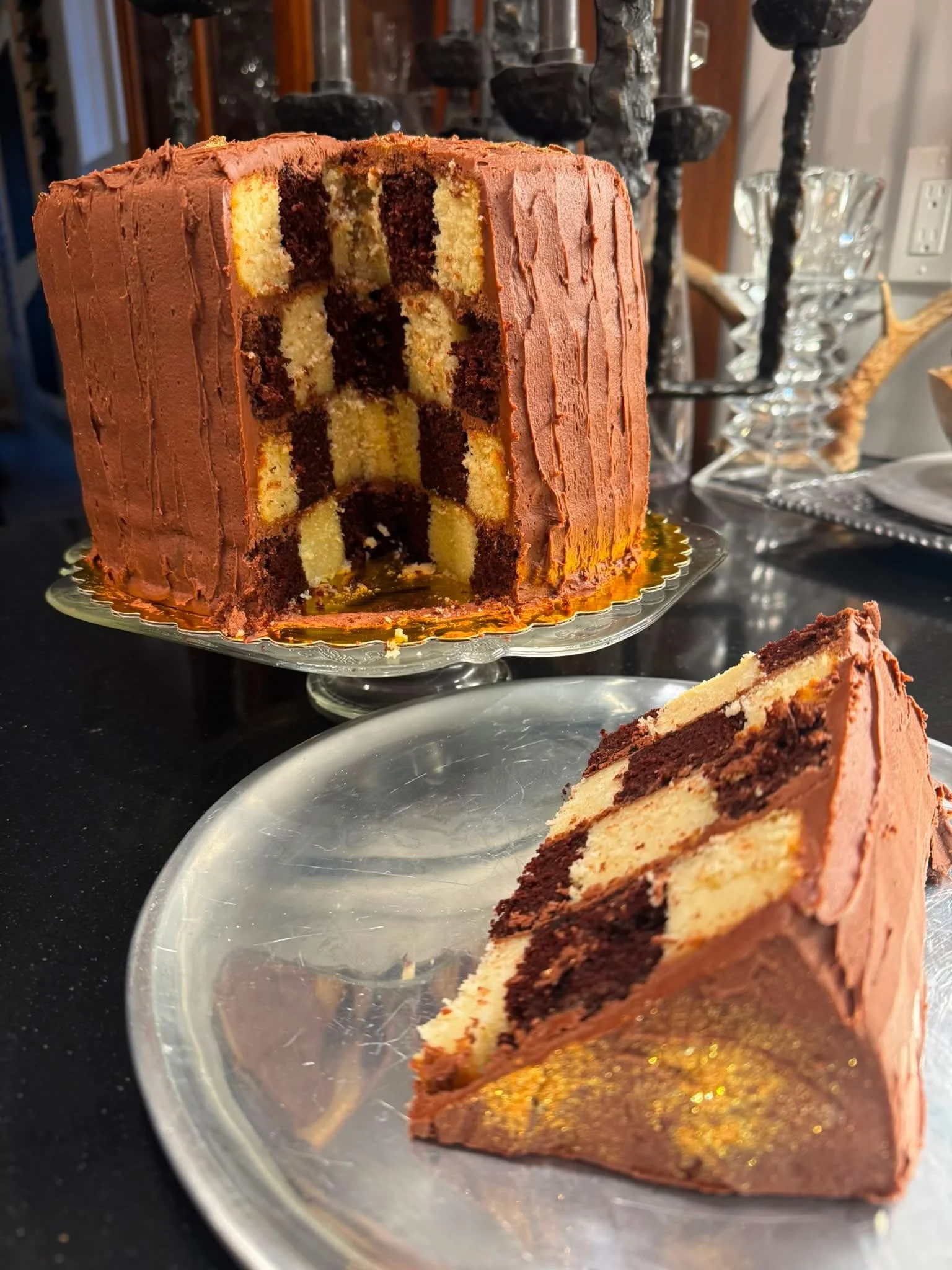 A tall layered cake with chocolate and vanilla sponge, decorated with chocolate frosting, is partially sliced on a glass cake stand. A slice of the cake is served on a plate in front.