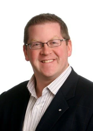 Portrait of a smiling man with glasses, wearing a dark blazer and a white dress shirt against a white background.
