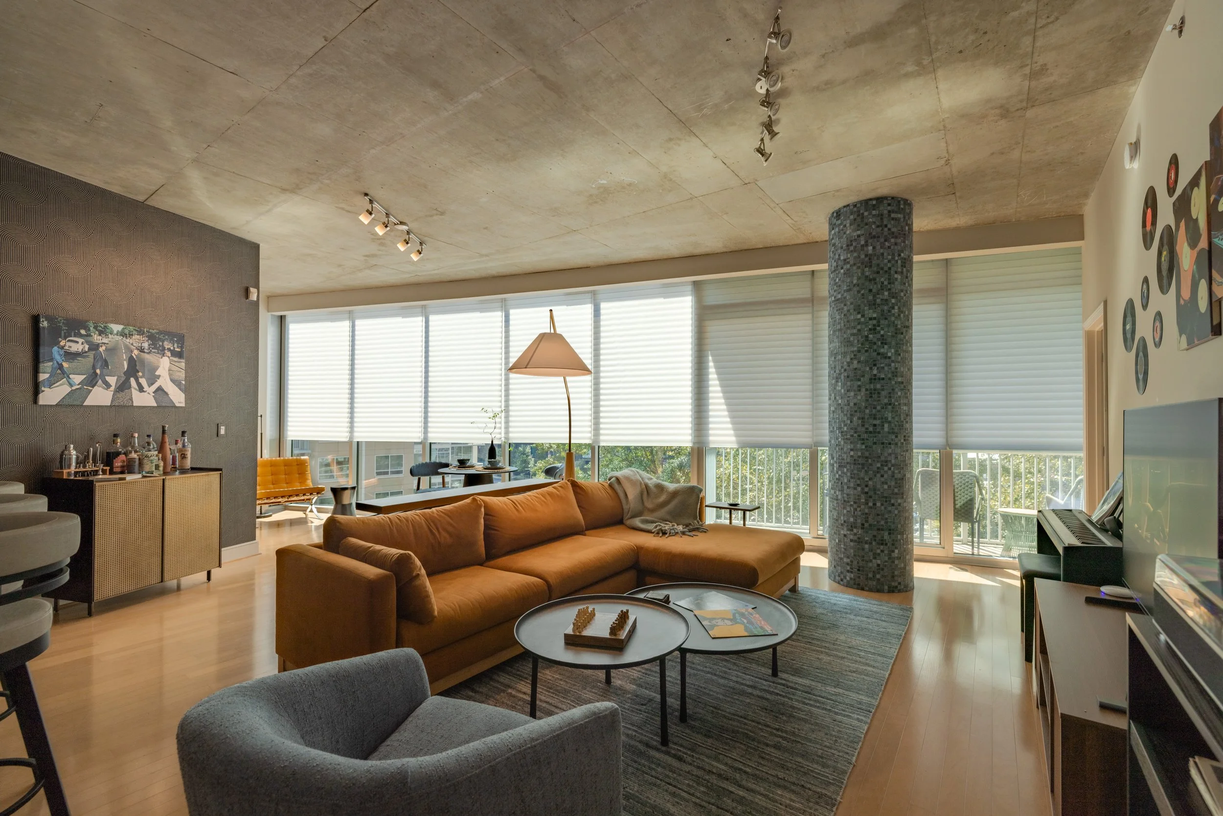 Modern living room with large windows, beige sofa, gray armchair, black coffee table, and a curved balcony with railing. Decor includes artwork on the wall, a floor lamp, and a mosaic column, with sunlight streaming through blinds.