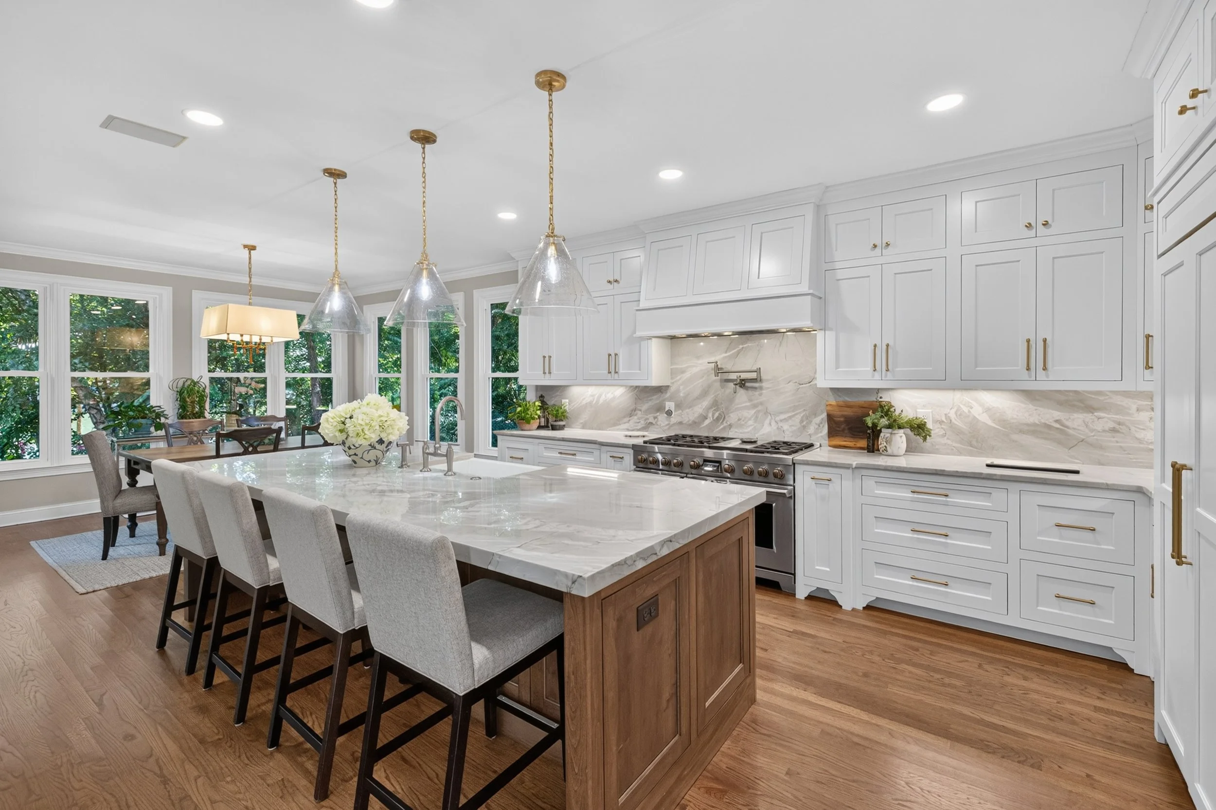 Modern kitchen with white cabinets, marble backsplash, wooden island, and pendant lights, with a dining area and large windows showing greenery outside.