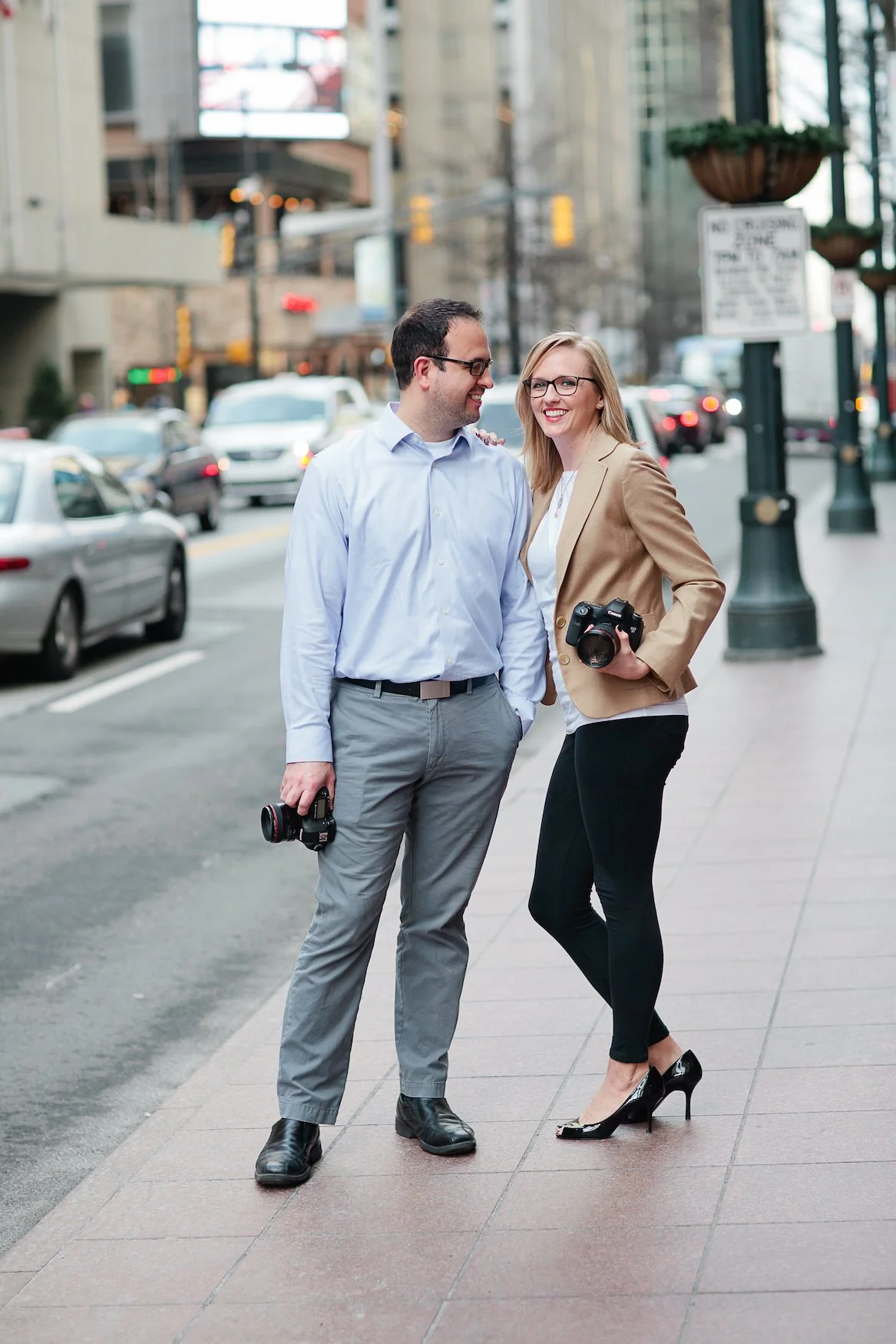 A man and woman stand on a city sidewalk, smiling at each other. The woman holds a camera, and the man holds another camera in his hand. Cars are parked along the street behind them, and tall buildings line the background.