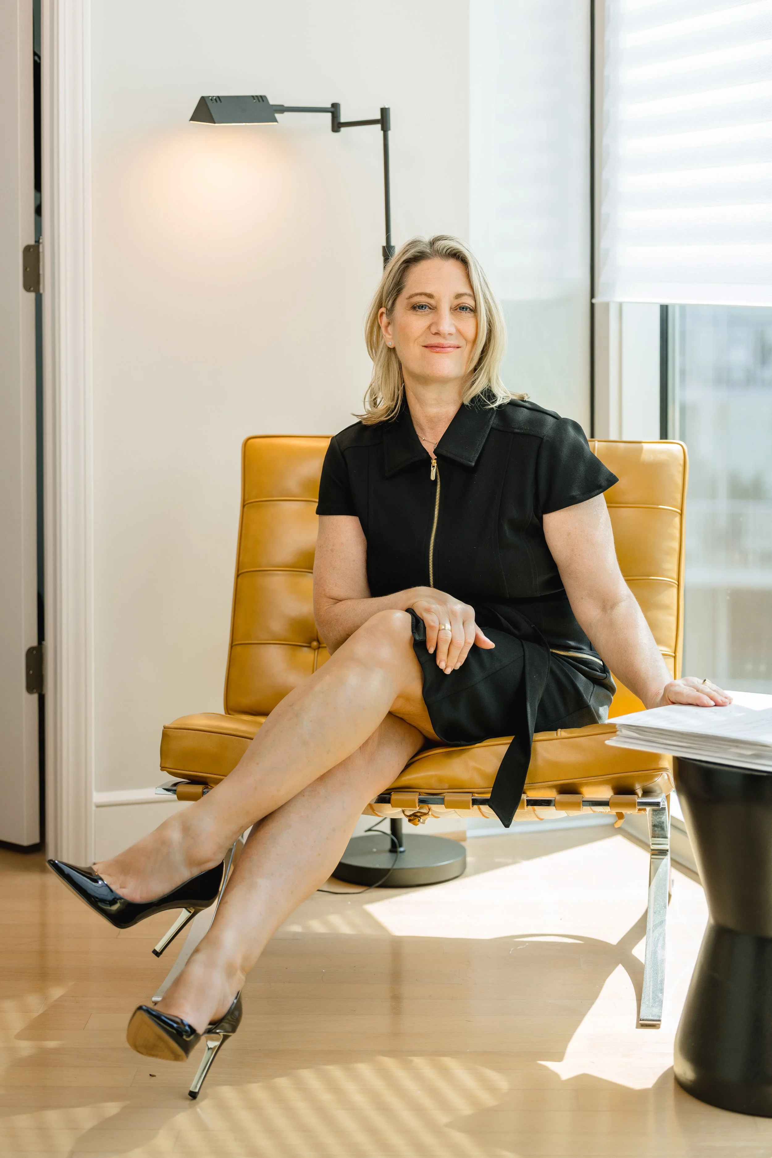 A woman sitting on a tan chair with her legs crossed, wearing a black dress and black high heels, in an office setting with a window and a modern lamp behind her.