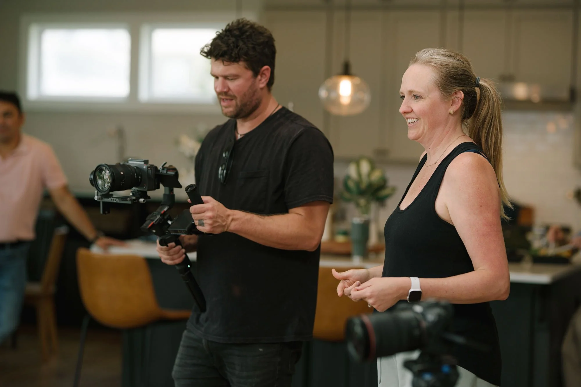 A man holding a camera stabilizer and a woman smiling, standing in a well-lit indoor space with kitchen cabinets in the background.