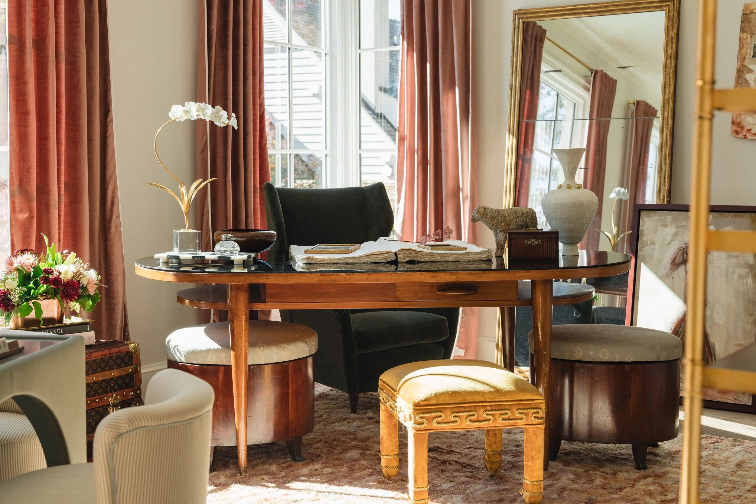 A cozy dining room with a wooden table surrounded by a black upholstered chair and a cushioned stool. The table has an open book, a small decorative box, and vases with flowers. Large windows with pink curtains let in natural light, and there are dec