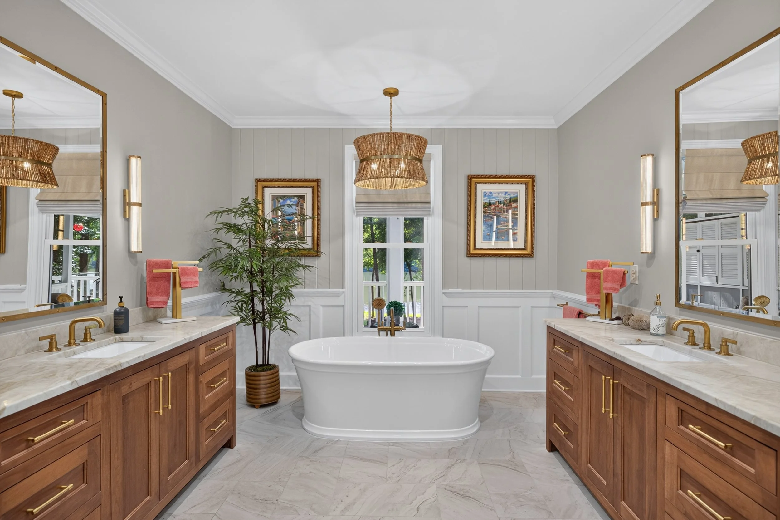 A spacious bathroom with a central free-standing bathtub beneath three large windows. There are two wooden vanities with marble countertops, gold fixtures, and pink towels. Two large mirrors with gold frames and modern wall sconces are mounted above 