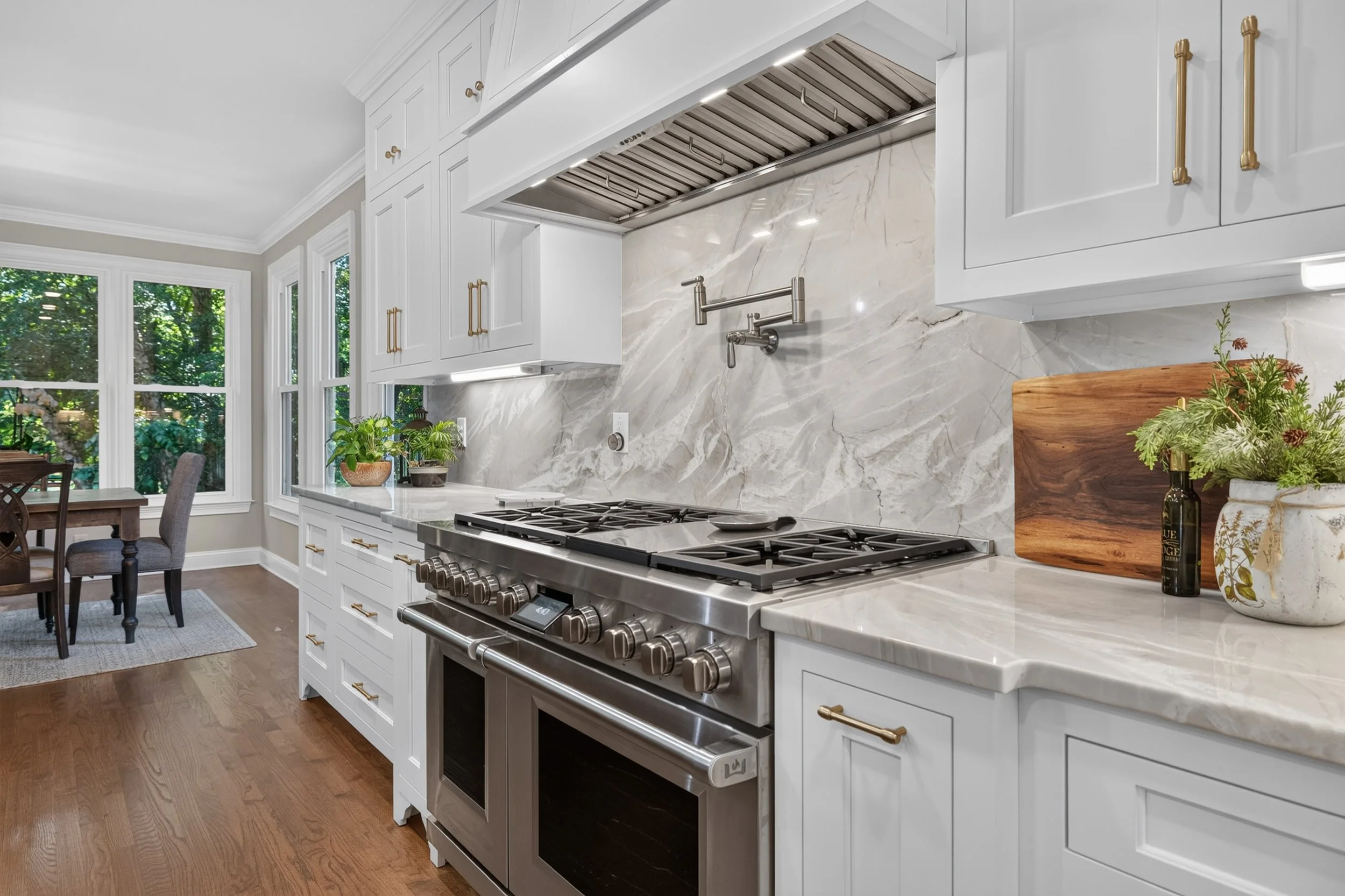 A modern kitchen with white cabinets, a marble backsplash, stainless steel stove, and wooden accents. There are two plants on the counter and a wooden cutting board. Large windows reveal greenery outside.