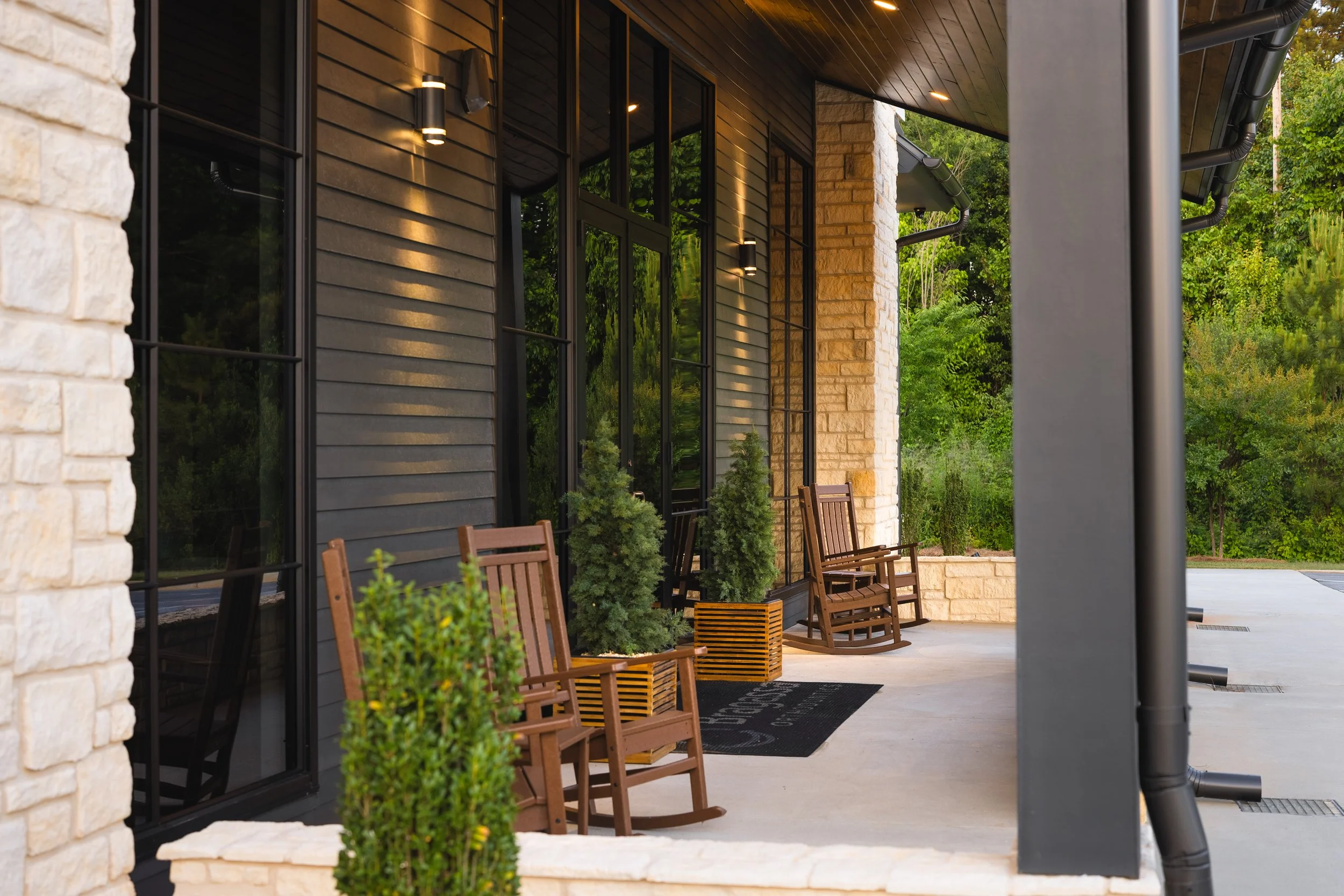 Front porch of a modern house with black wood siding, windows, outdoor seating, potted plants, and a black mat.