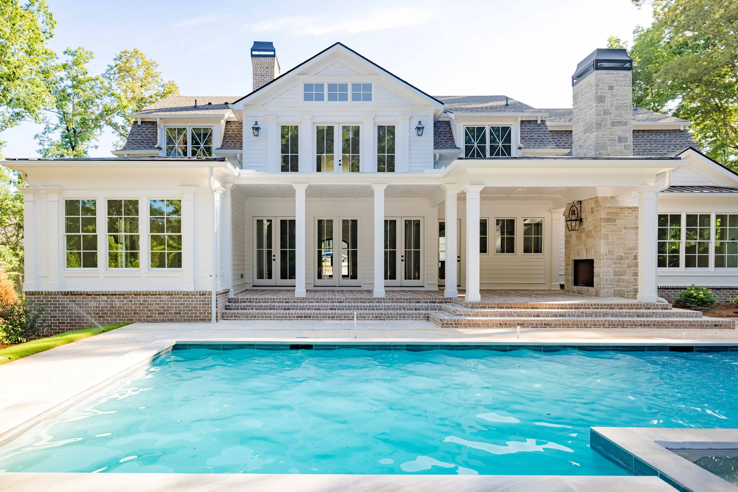 Backyard view of a large white house with multiple windows, a chimney, and a covered patio area with steps leading down, overlooking a swimming pool in the foreground.
