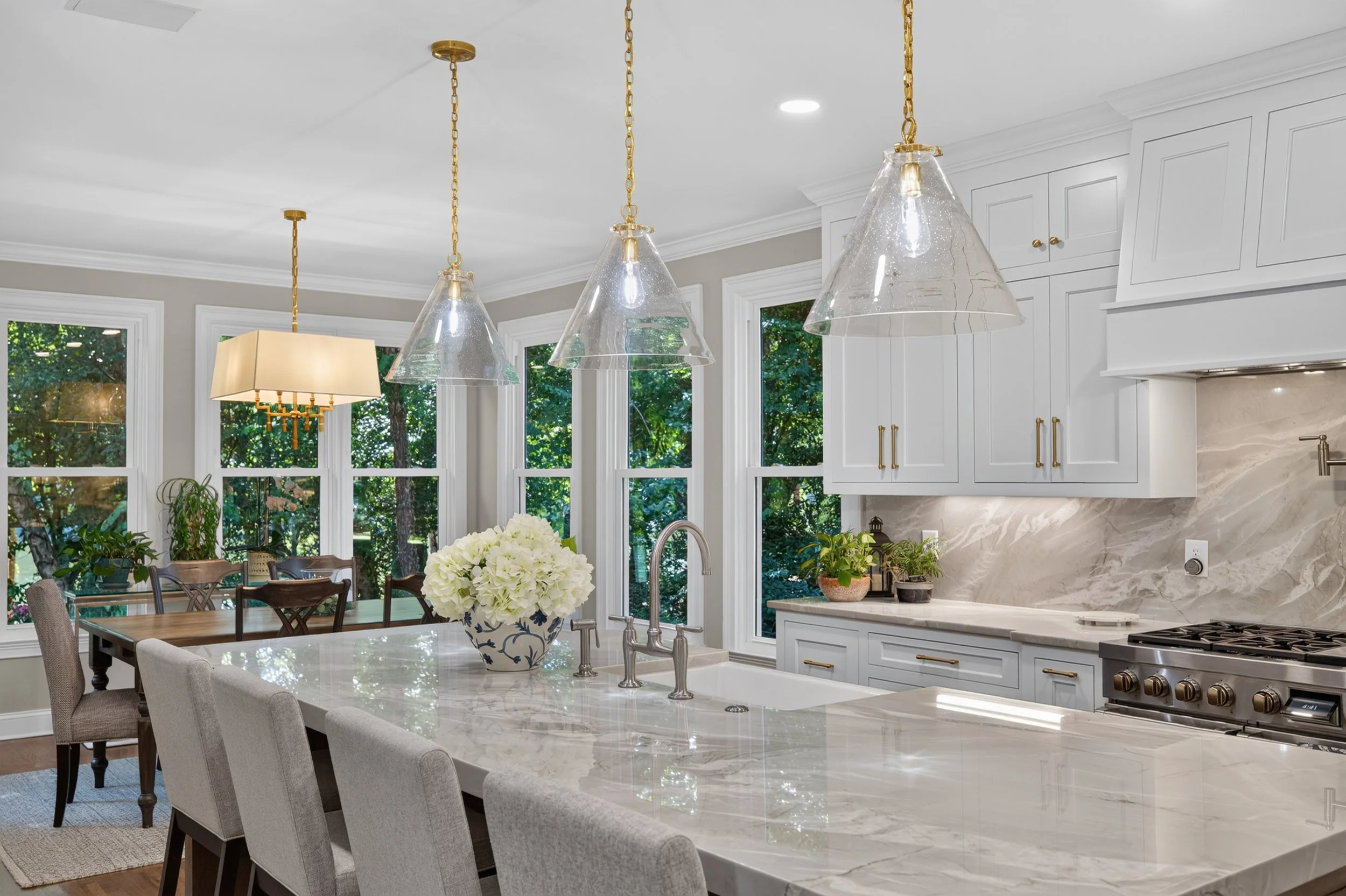 Modern kitchen with white cabinets, marble countertops, and large windows showing green trees outside. Three glass pendant lights hang above a kitchen island, which has a vase with white flowers. A dining table with chairs is visible in the backgroun