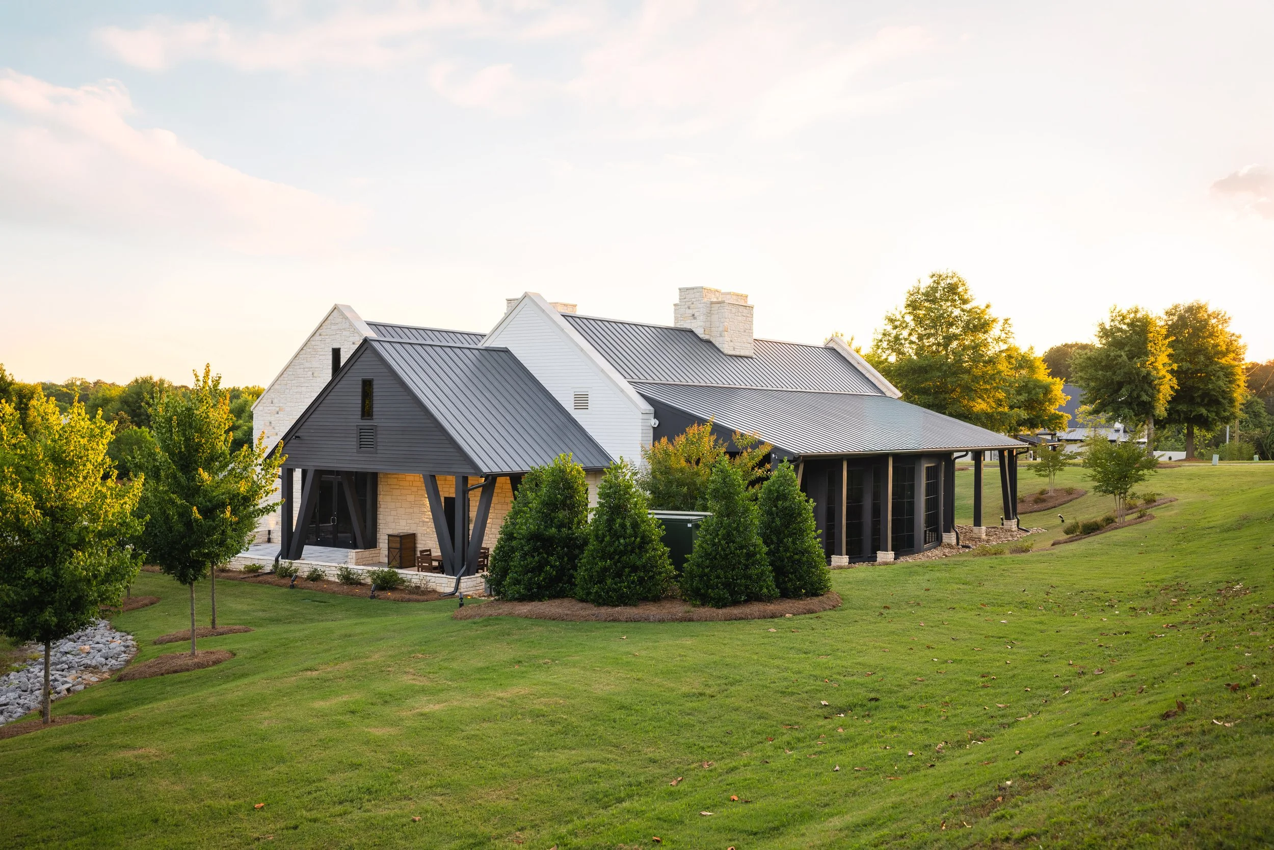 Large modern house with a gray metal roof, white brick walls, black accents, surrounded by well-maintained grass and various trees, during sunset.