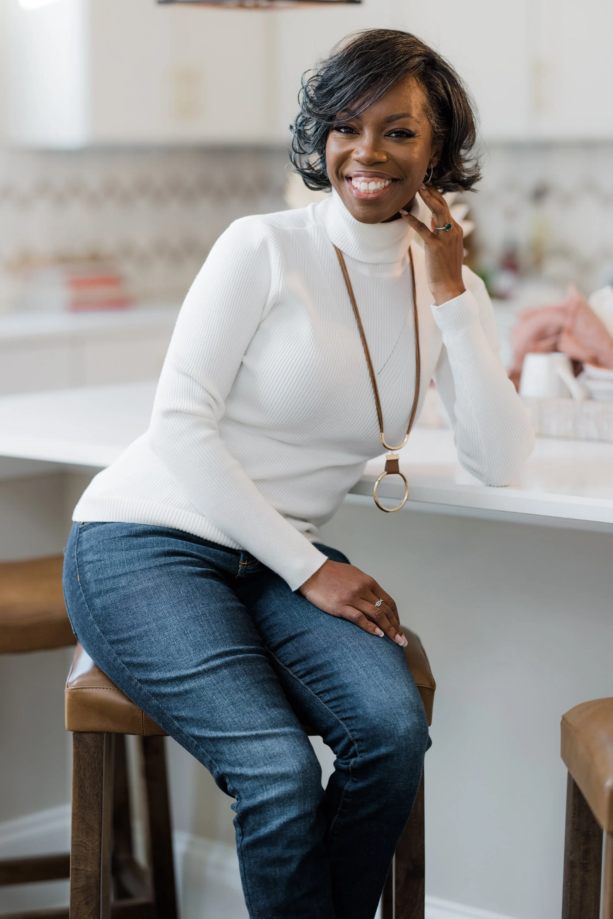 A woman with dark, curled hair and a white turtleneck sweater sitting on a bar stool in a kitchen, smiling at the camera, with her hand on her neck and wearing a ring and necklace.