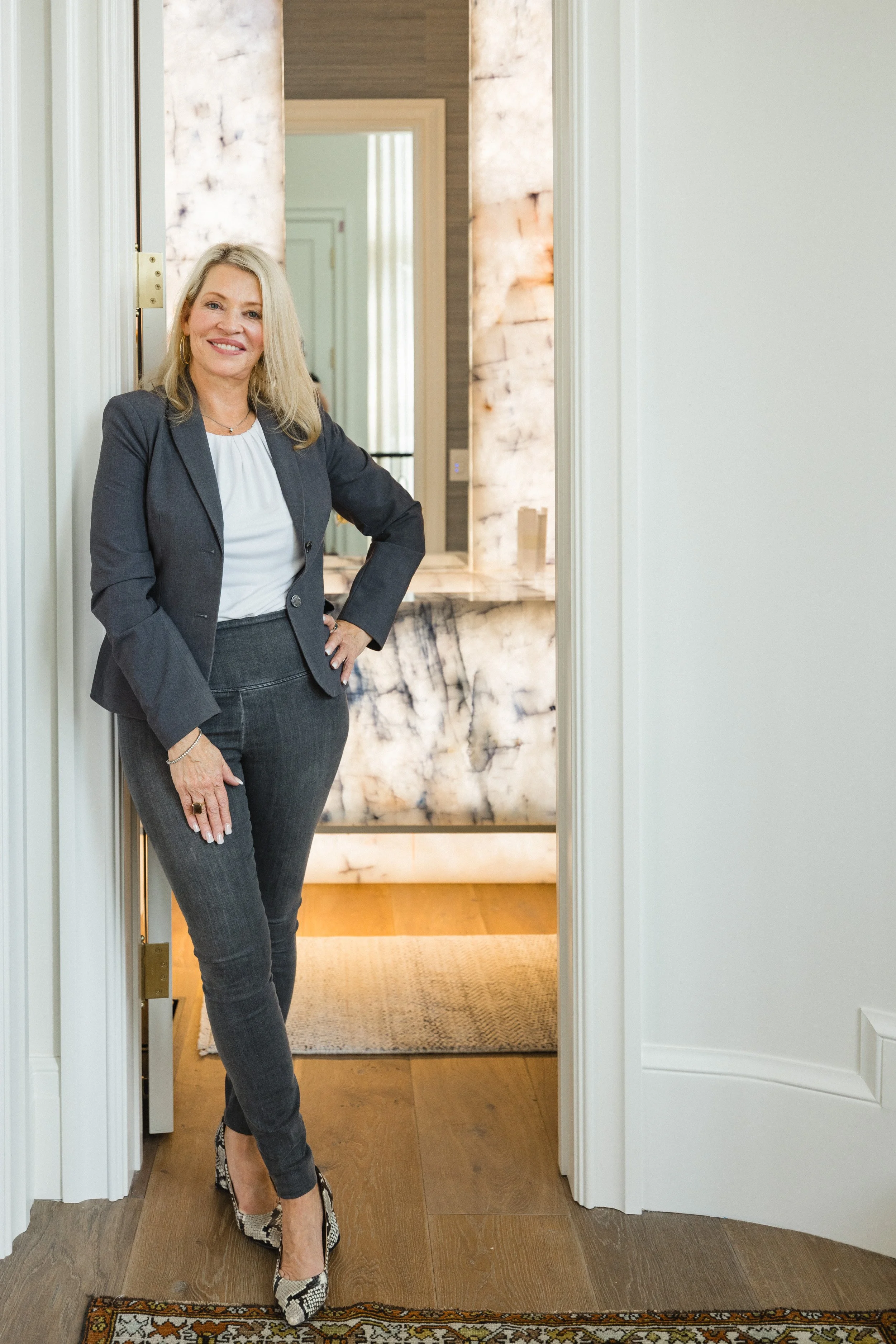 A woman in a gray suit and snakeskin heels standing in a doorway smiling at the camera in a well-lit room with marble and wood decor.