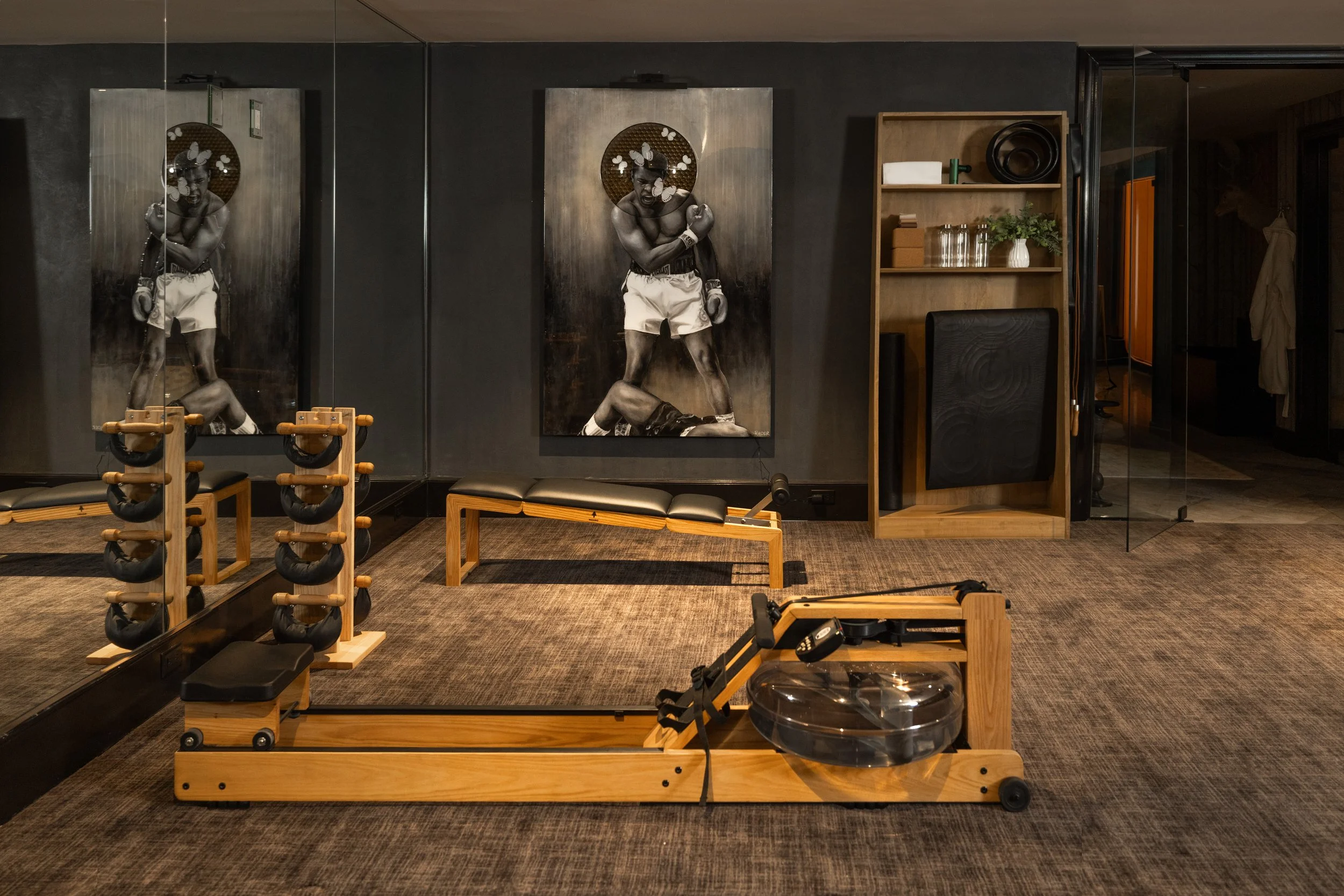 A room with boxing equipment, including wooden and padded benches, a water-filled boxing training punching bag, and large black and white photographs of a boxer on the wall. The room has dark walls, mirrors, and a wooden shelf with plants and accesso