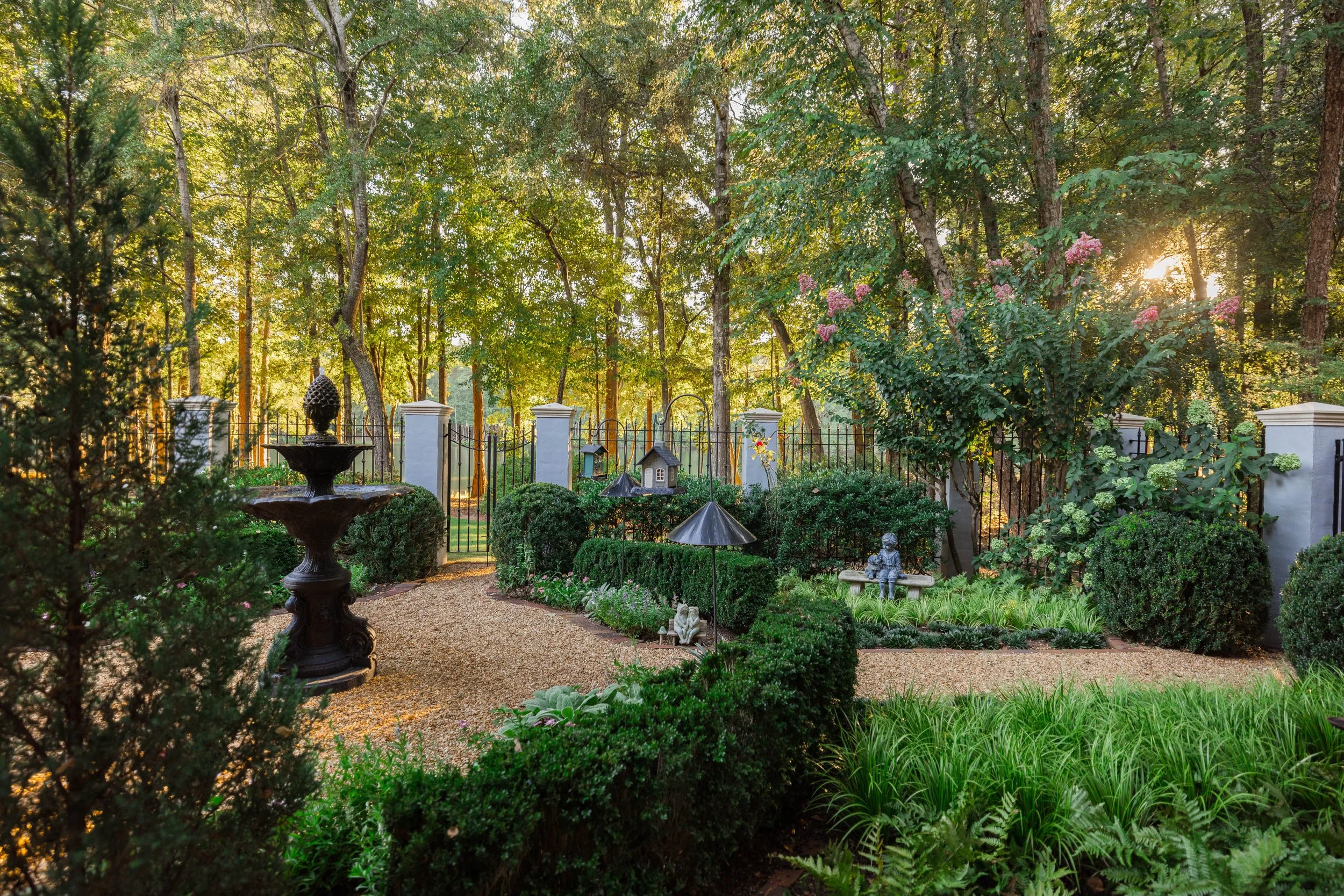 A lush garden with tall trees, a fountain, statues, and a black iron fence, illuminated by sunlight filtering through the leaves.