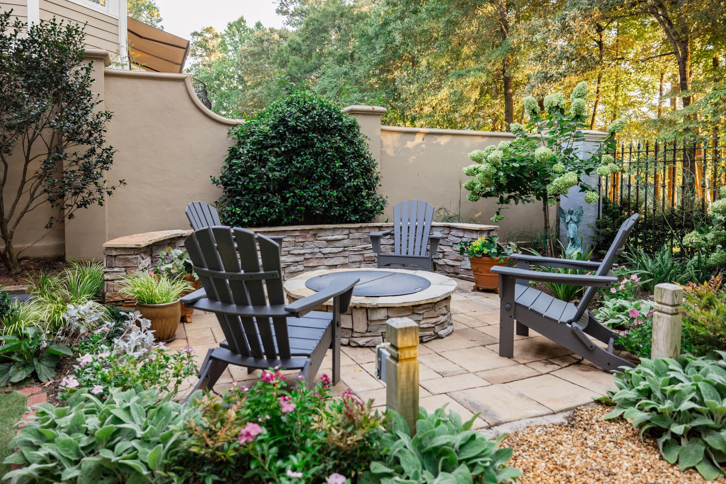 A backyard patio area with potted plants, Adirondack chairs, a stone fire pit, and a beige stucco wall, surrounded by trees and greenery.