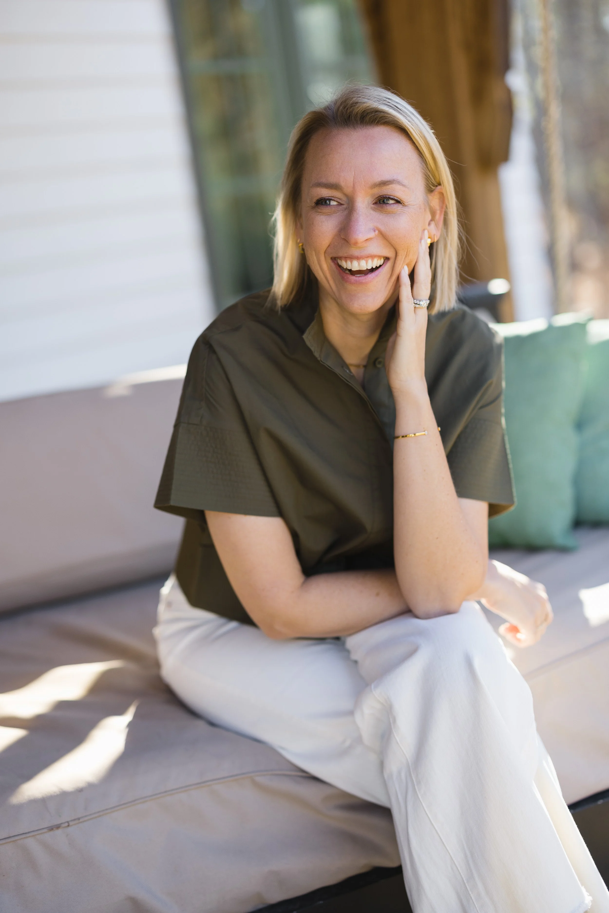 A woman with blonde hair smiling, sitting on an outdoor sofa with a white shirt and beige pants, touching her face with her right hand.