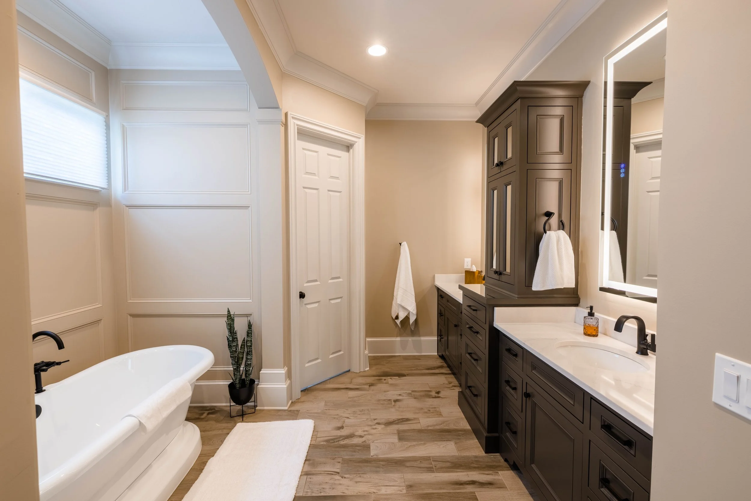 A modern bathroom with a clawfoot bathtub, a dark gray vanity with a white marble countertop, a large mirror with built-in lighting, wooden flooring, and a small potted plant.