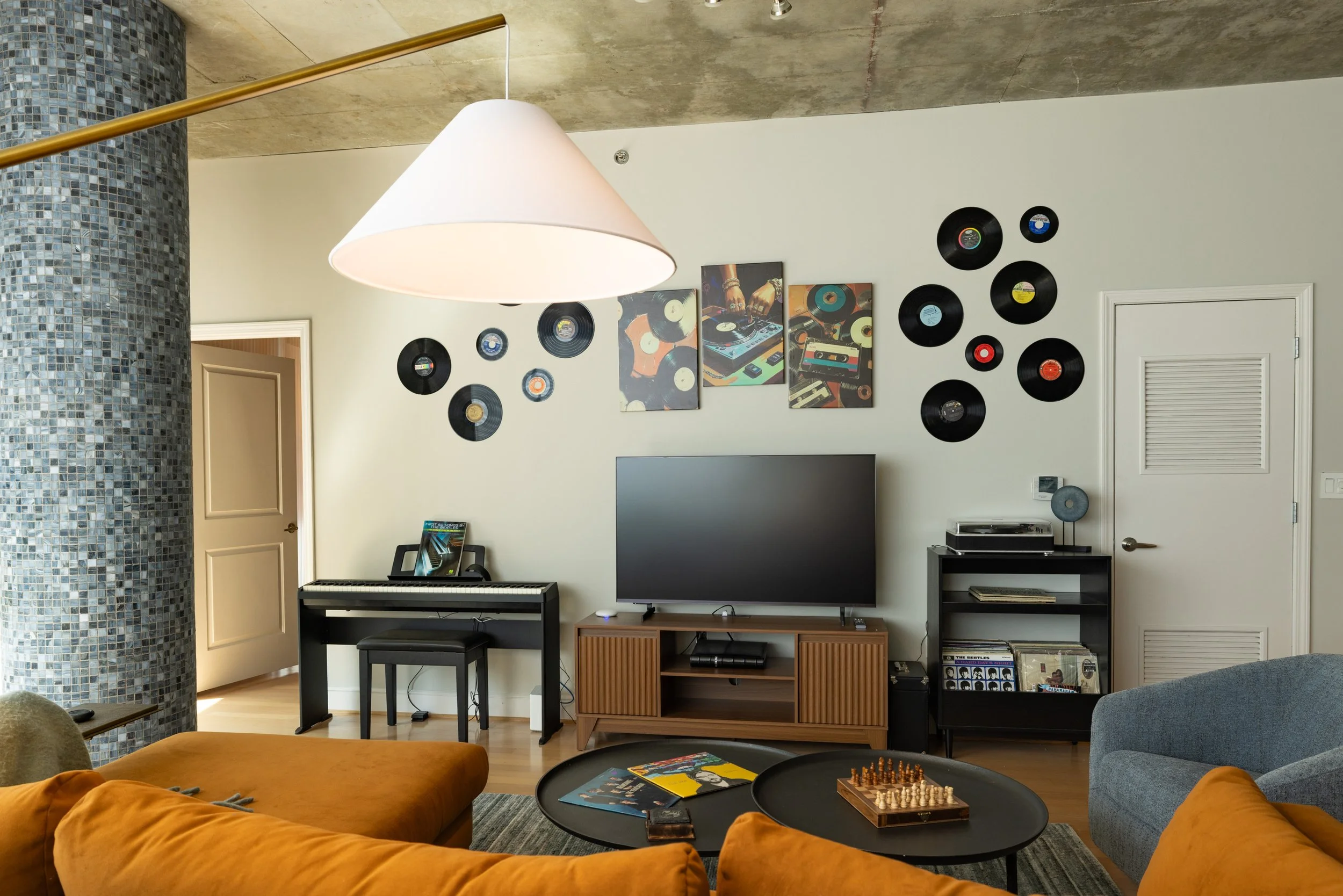 Living room with a white wall, decorated with vinyl records and colorful artwork, a television, a piano, and a black side table with records. A large white pendant lamp hangs from the ceiling, and there is a mosaic column on the left.