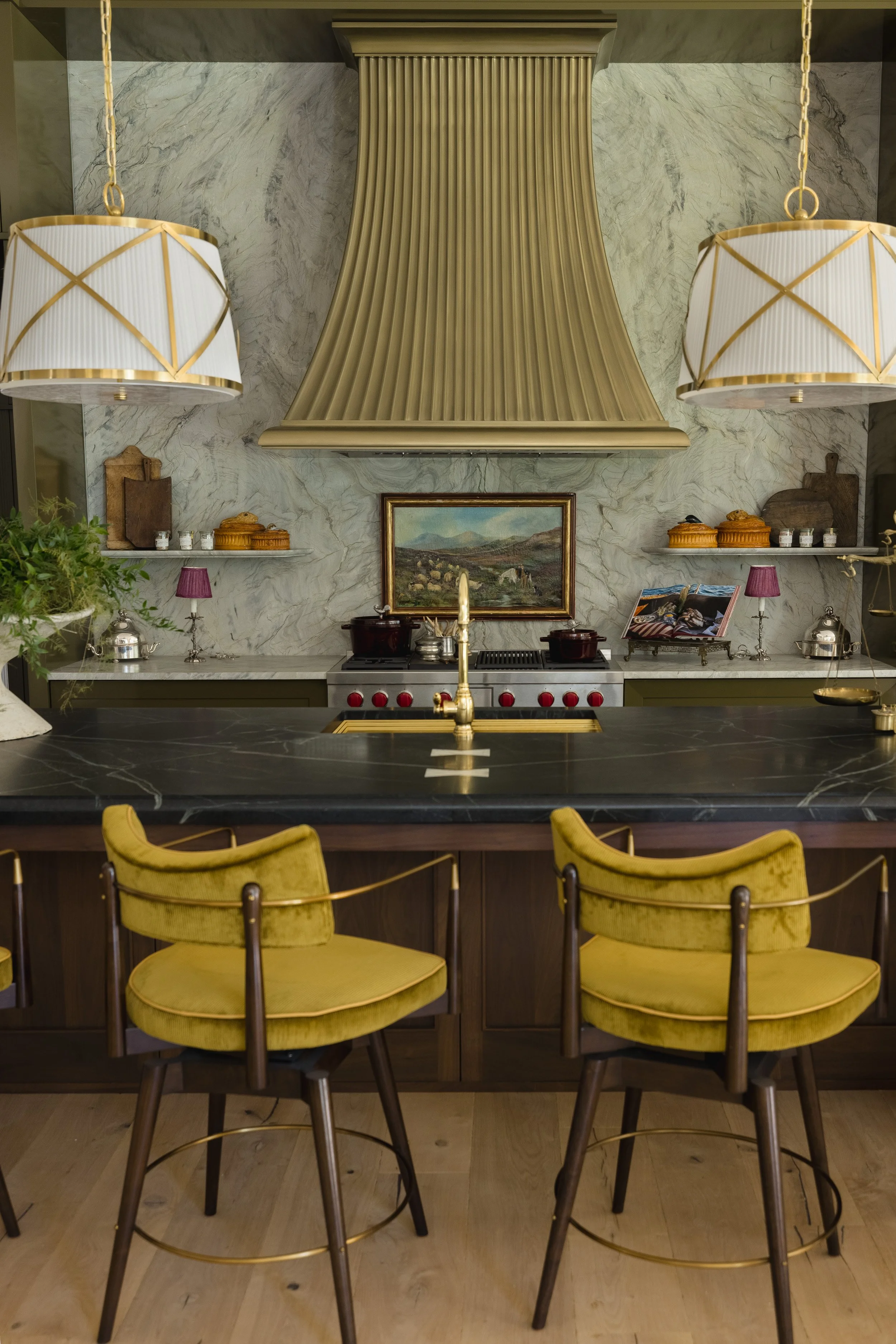 A modern kitchen with a black marble countertop, yellow velvet chairs around a black island, a marble backsplash, gold accents, and two hanging white and gold lamps.