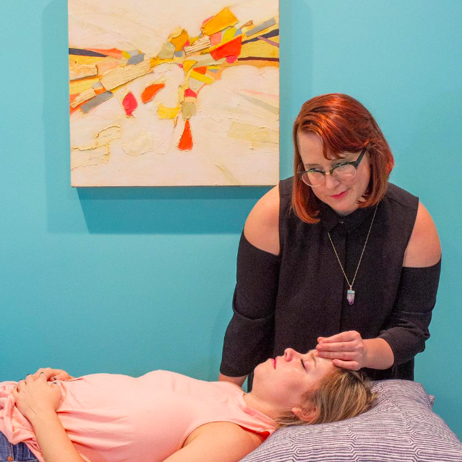A woman lying on a massage table with her eyes closed and hands on her stomach, receiving a facial or head massage from another woman with glasses, in a room with blue walls and abstract painting.