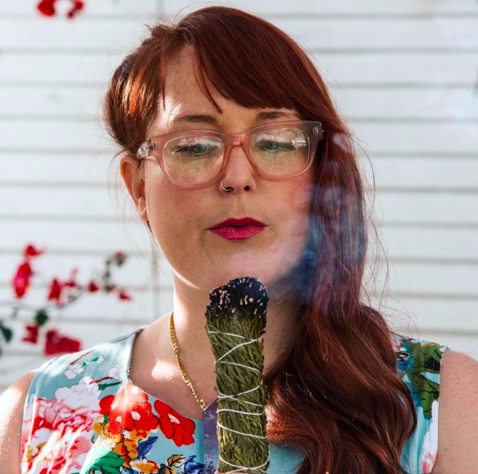 A woman with red hair and glasses exhales smoke from a smoked sage bundle outdoors, with a white wall and some flowers in the background.