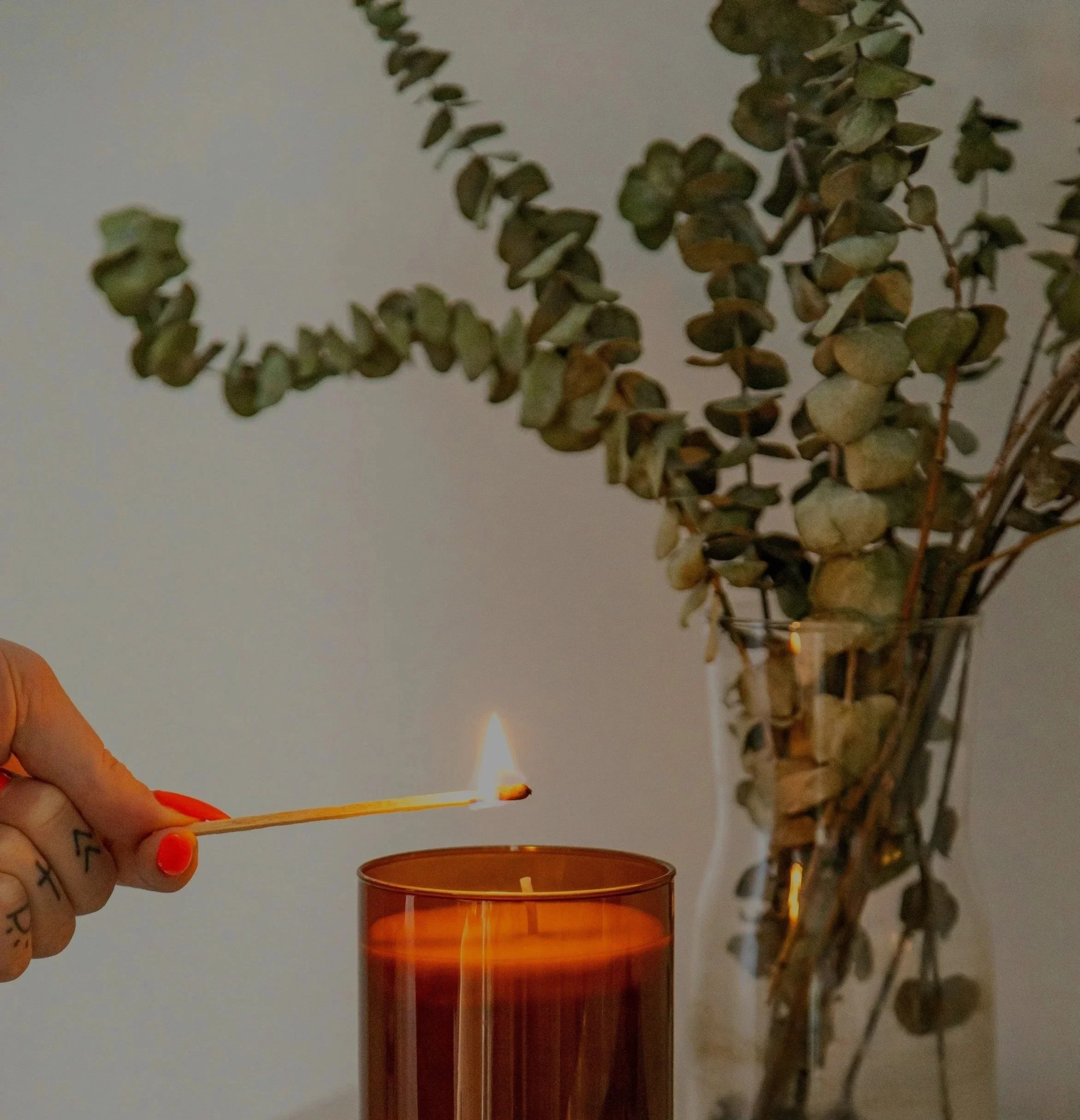 Person holding match lighting a candle in a glass jar with dried eucalyptus branches in the background.