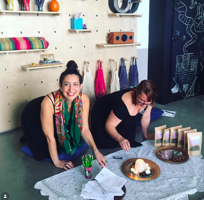 Two women are kneeling on the floor, smiling and engaging in a religious or ceremonial activity on a white lace tablecloth. One woman has dark hair in a bun, wearing a black dress and colorful scarf, while the other has brown hair, glasses, and is dressed in black. There are papers, small bags, candles, and stones on the table, with a dark wall featuring chalk drawings of a snake and other shapes behind them.