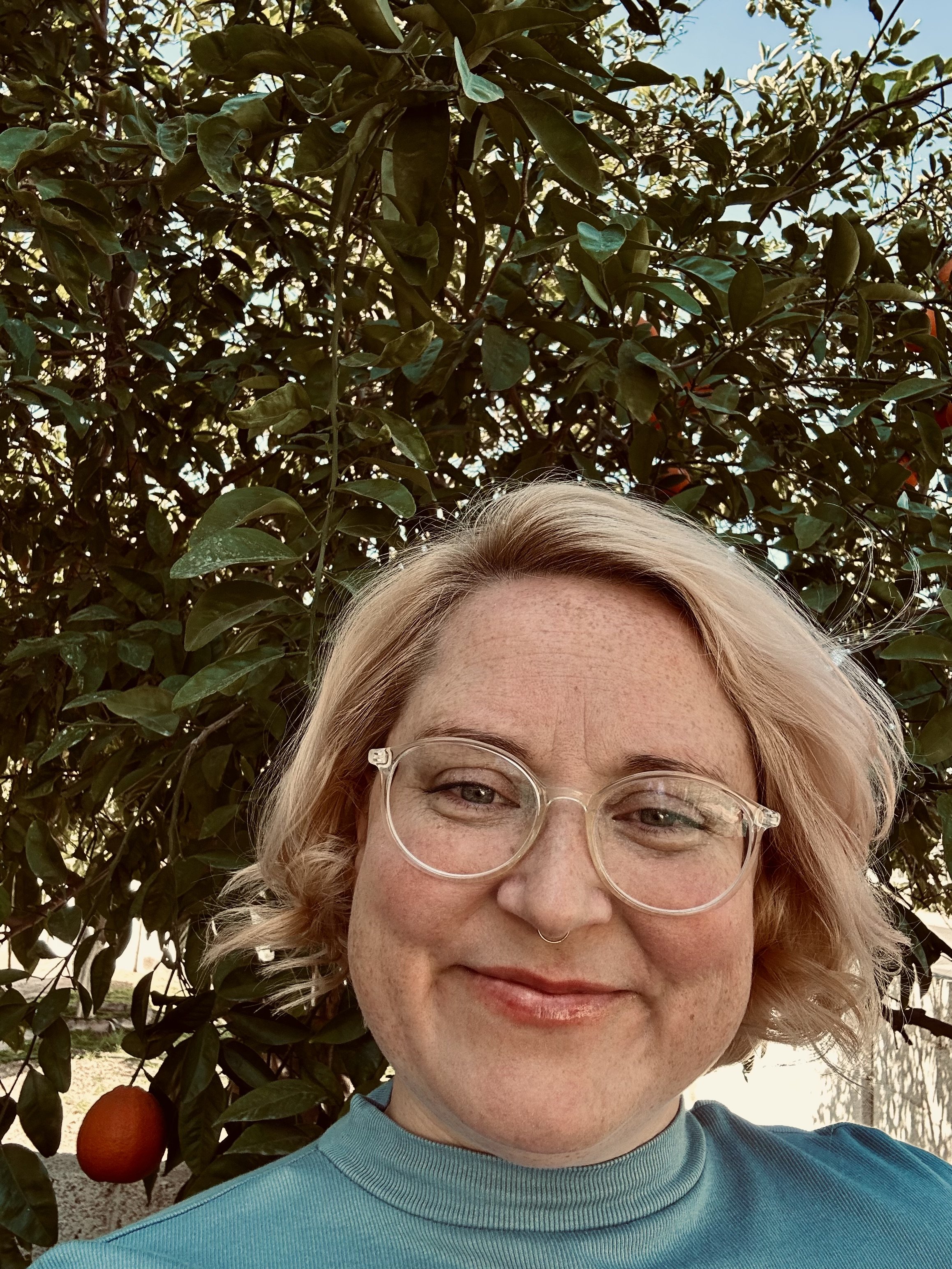 A woman with blonde hair and glasses smiling outdoors in front of a tree with orange fruit.