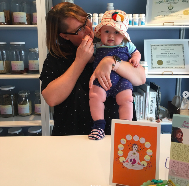 A woman holding a baby in a store or office environment, with shelves of jars and certificates in the background, and a colorful greeting card on the counter.