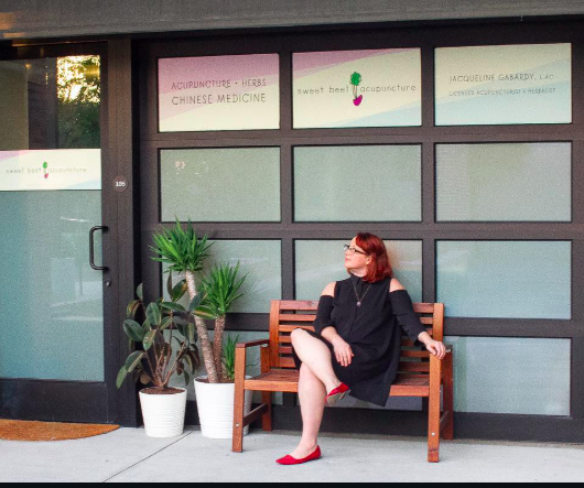 A woman with red hair, wearing glasses and a black dress, sits on a wooden bench outside a storefront with glass panels. There are potted plants beside her, and signs above the window advertising acupuncture, herbs, Chinese medicine, and a sweet beet. A door to the left has a black handle.