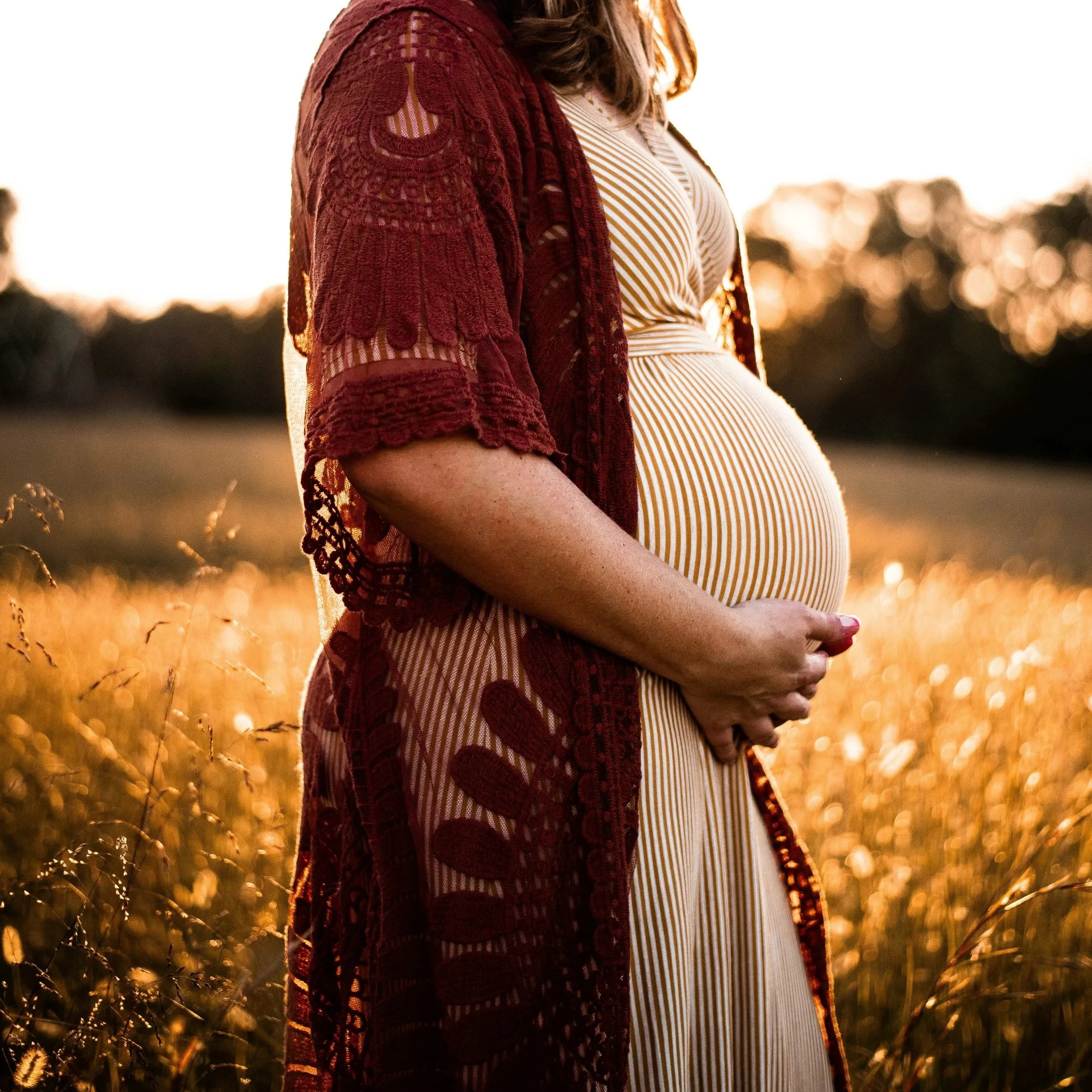 A pregnant woman wearing a white and beige striped dress and a maroon lace cardigan, standing outdoors in a field of tall grass during sunset.