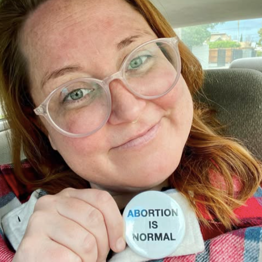 A woman with red hair and glasses smiling while holding a button that says 'Abortion is Normal' in a car.