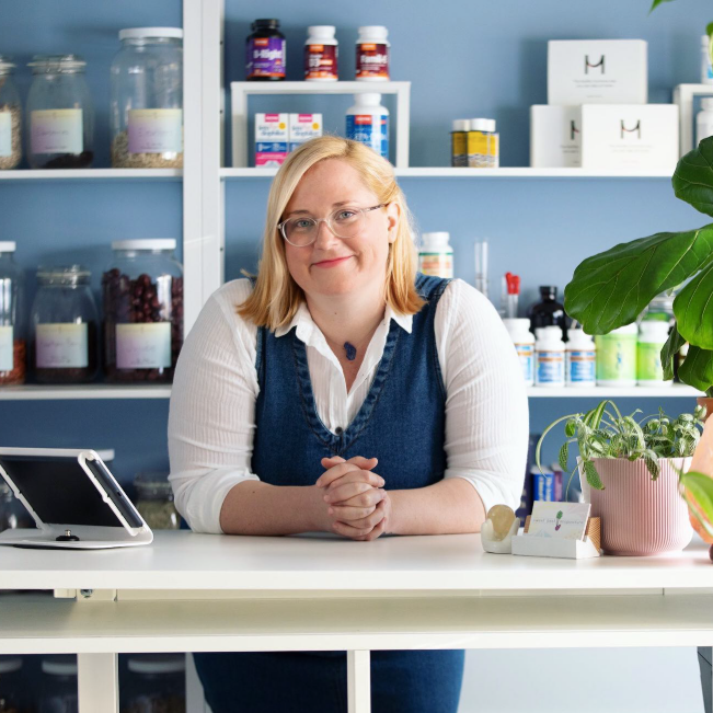 A woman with blonde hair, glasses, and a white shirt under a denim vest sits at a white counter with her hands clasped. She is smiling and in a pharmacy or supplement store with shelves of bottles and boxes behind her, and a potted plant on the right side of the counter.