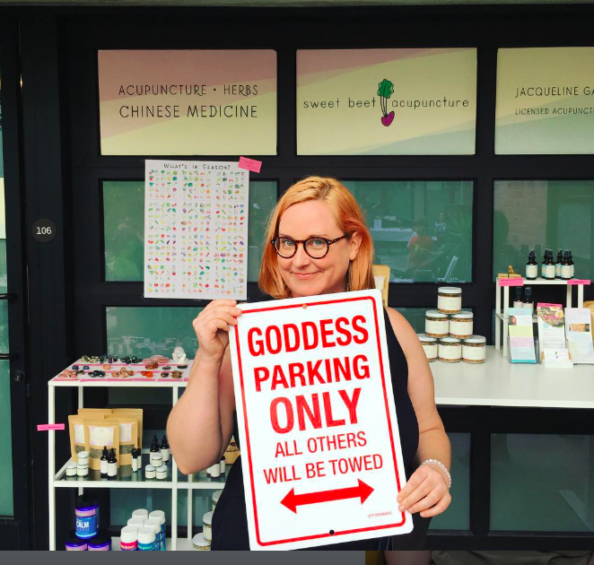 A woman with glasses holding a 'GODDESS PARKING ONLY' sign in a store that sells acupuncture and herbal products.