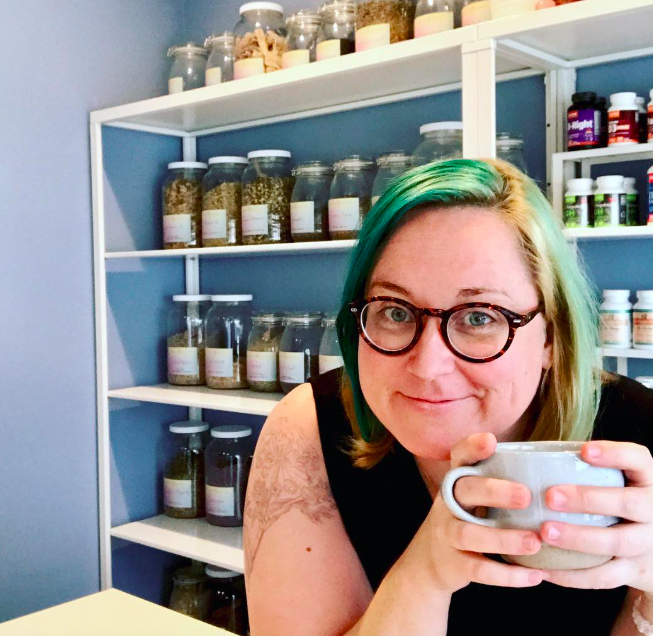 Woman with dyed green hair and glasses holding a mug, seated in front of shelves filled with jars of herbs or teas in a room with light blue walls.
