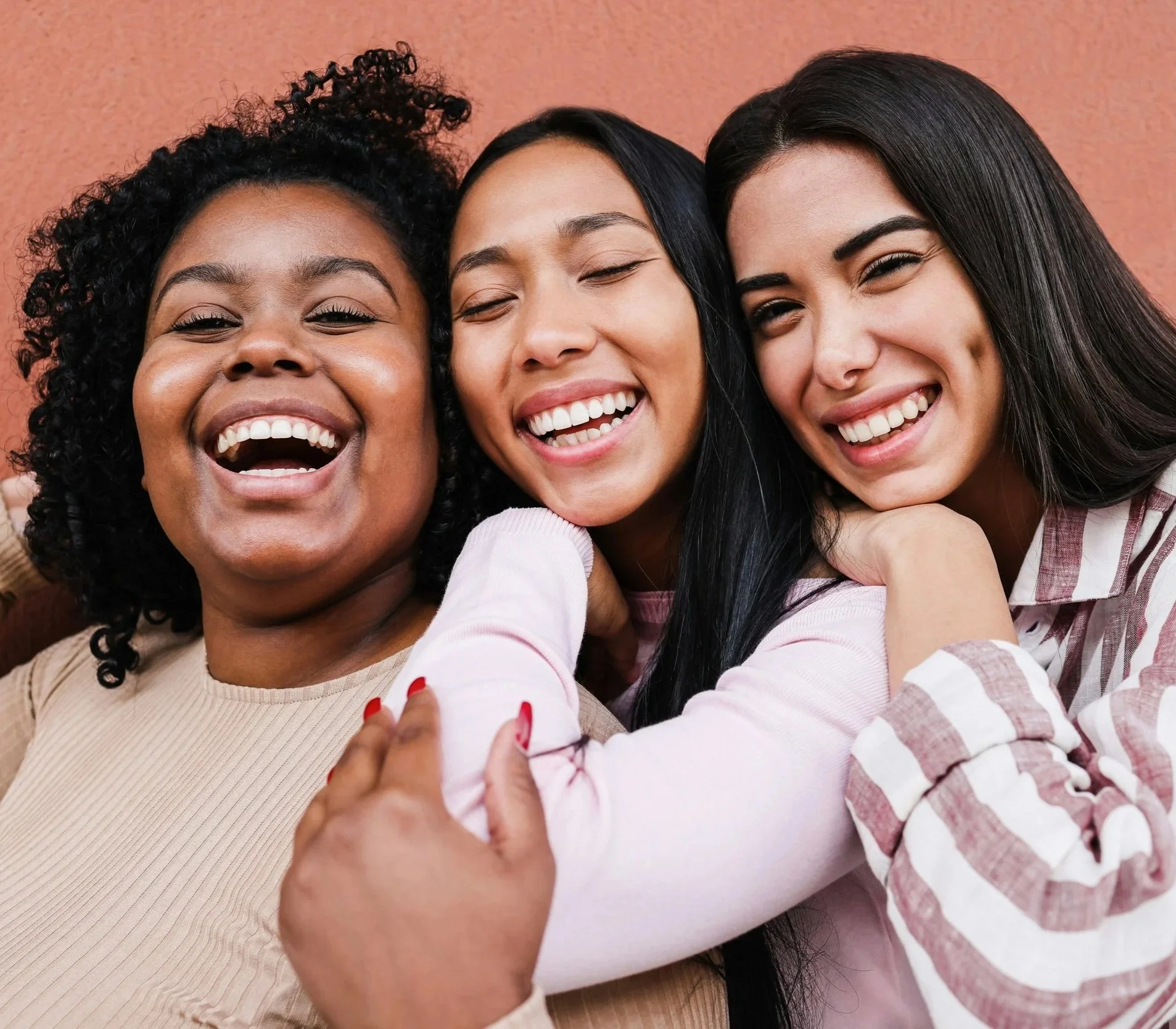 Three women of different ethnicities smiling and hugging each other, close-up photo with beige background.