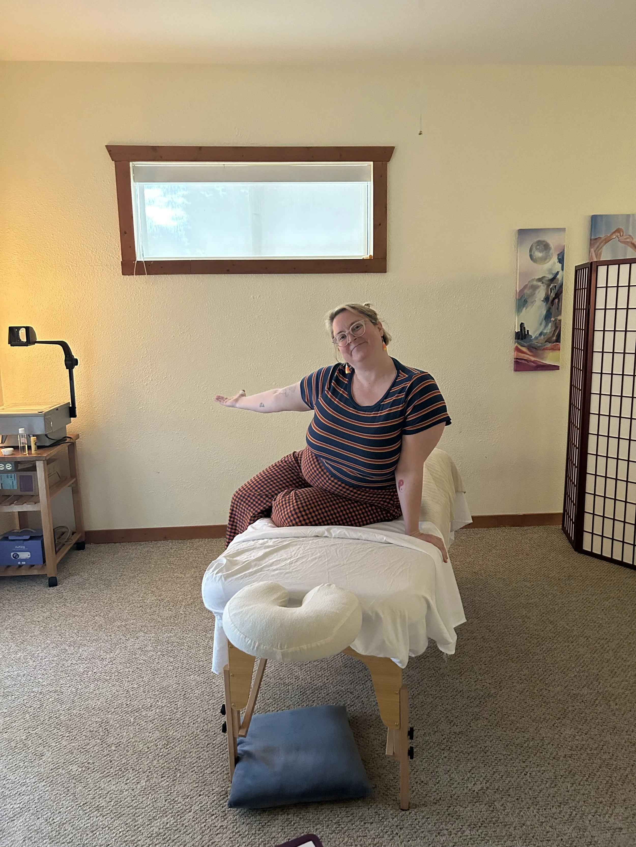 Person sitting on a massage table in a room, with their arm extended, as if receiving a treatment; decorations on the wall and a privacy screen in the background.