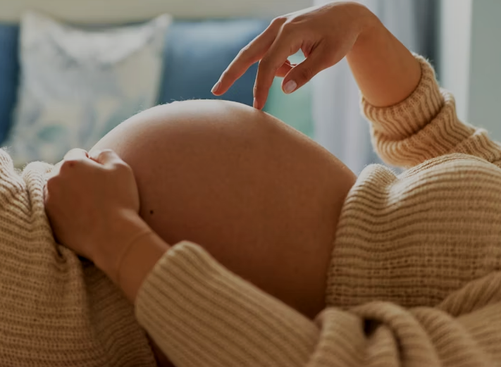 Close-up of a pregnant woman touching her belly while lying down, wearing a cozy beige sweater in a bright room.