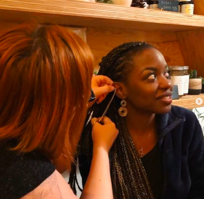 A woman with long braided hair is getting her ear pierced by another woman with red hair in a cozy, wooden setting, possibly a shop or cafe.