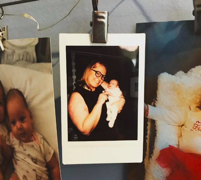 Photograph of a woman with glasses holding a baby, hanging on a clip against a wall, with parts of other photos visible around it.