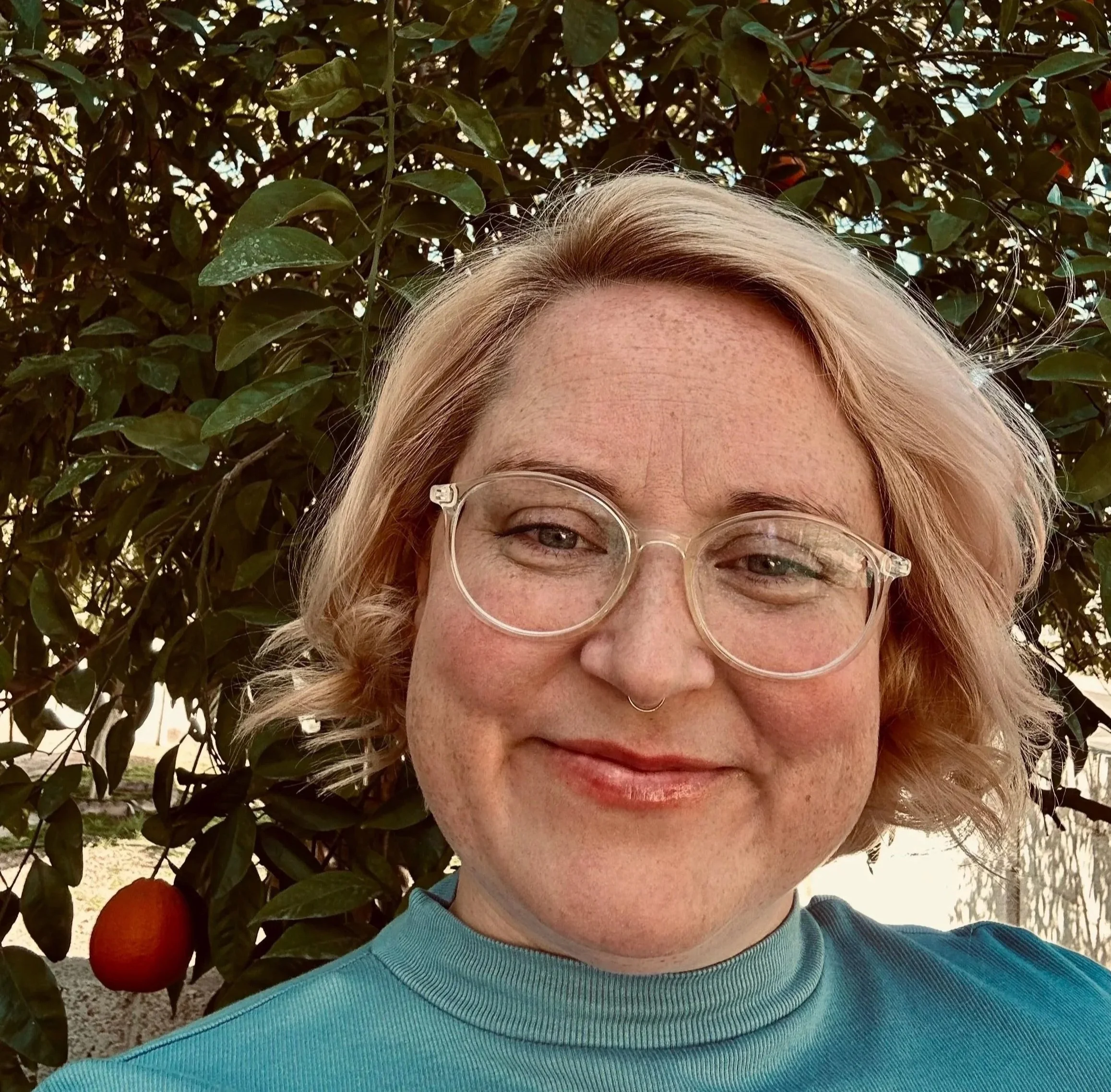 Part of a woman's face, including her forehead, eyes, and glasses, with a background of leafy green bushes and ripe red oranges in an outdoor setting.