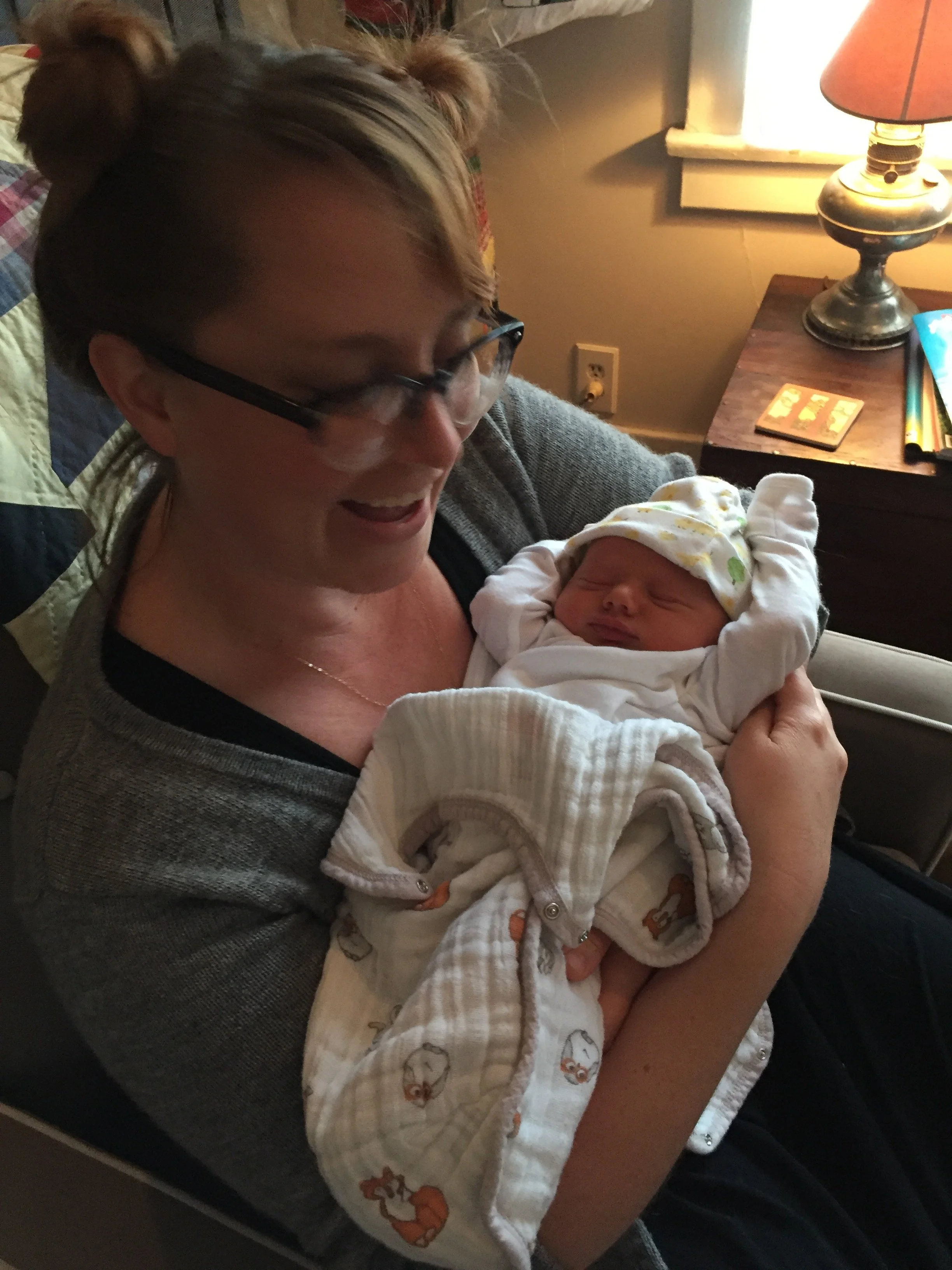 A woman is holding a newborn baby, smiling and looking at the baby. The baby is wearing a white hat and a cream-colored quilted outfit with animal designs, sleeping peacefully. They are sitting on a bed near a nightstand with a lamp, some magazines, and a small book in a room with a beige wall and a window.