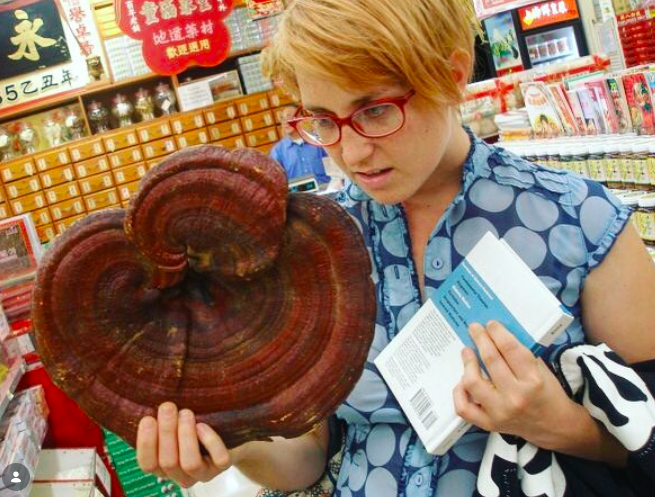 Woman with red glasses and blonde hair examining a large, multicolored mushroom in a grocery store aisle.