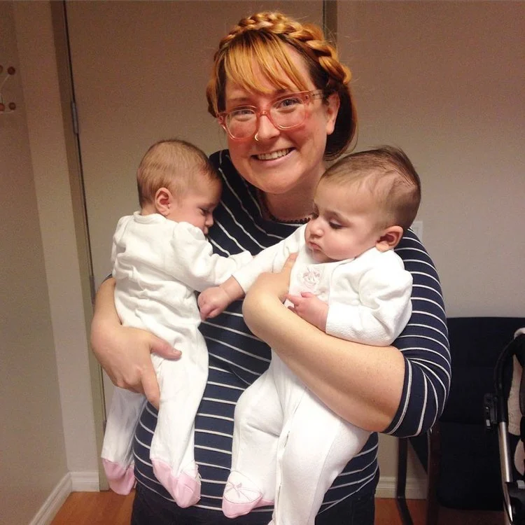 A smiling woman with glasses and braided hair holding two babies dressed in white.