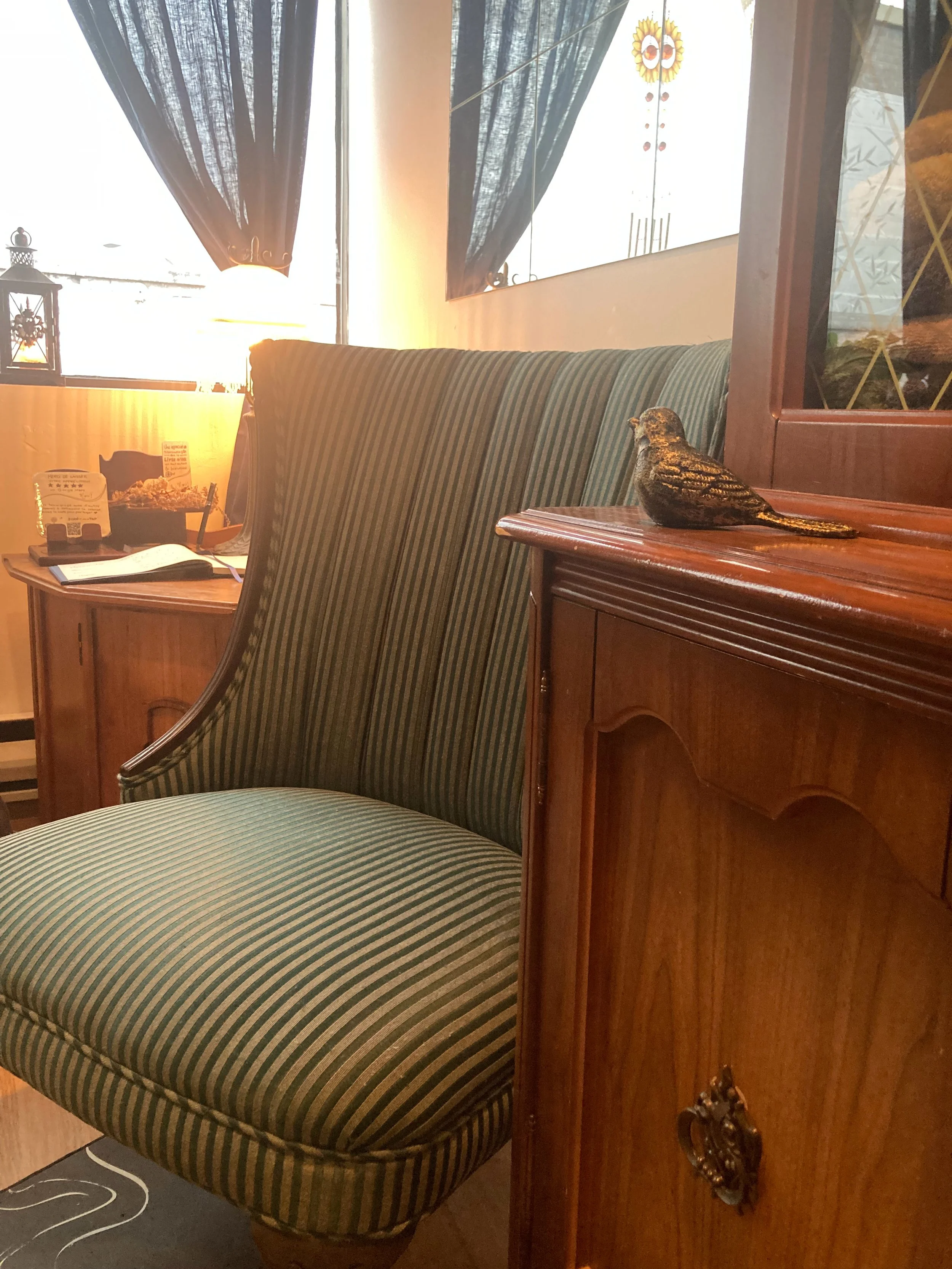 Intérieur avec des chaises à rayures vertes et beige, une table en bois avec une pièce décorative en forme d'oiseau, une fenêtre avec des rideaux noirs et une décoration murale représentant un soleil. Une lampe de table et une lanterne en verre sont visibles.