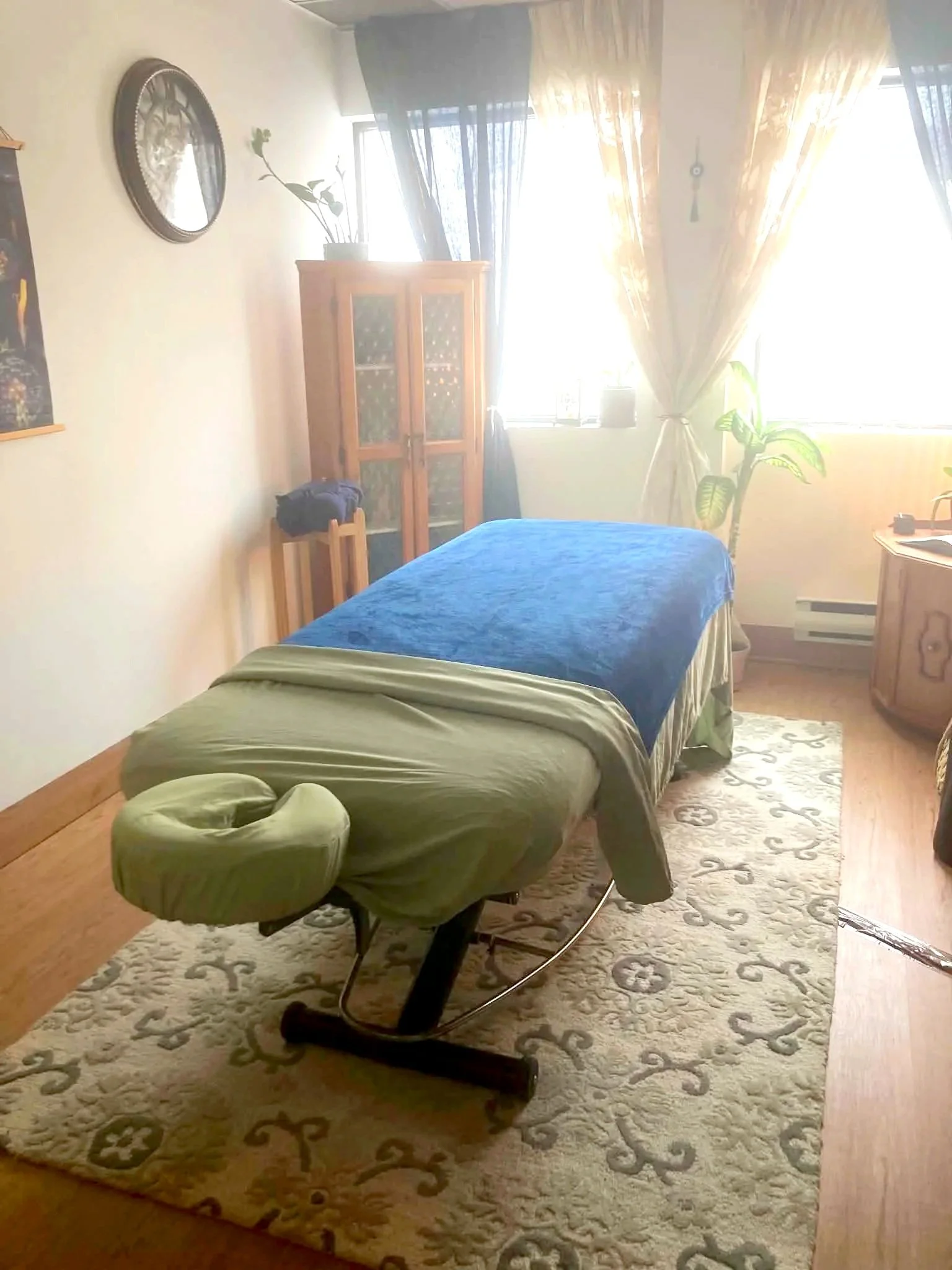 Une pièce lumineuse avec un lit, une commode en bois, une plante, un tapis beige avec motifs, des rideaux dorés et une table de massage au centre.