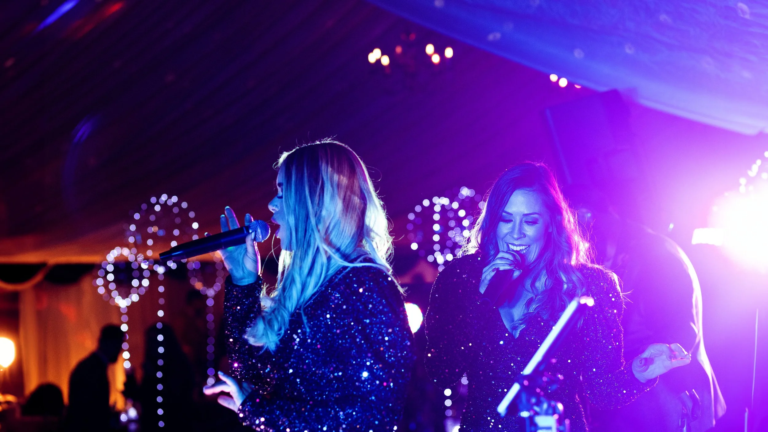Two women singing into microphones during a performance under colorful stage lights, with decorative strings of lights in the background.